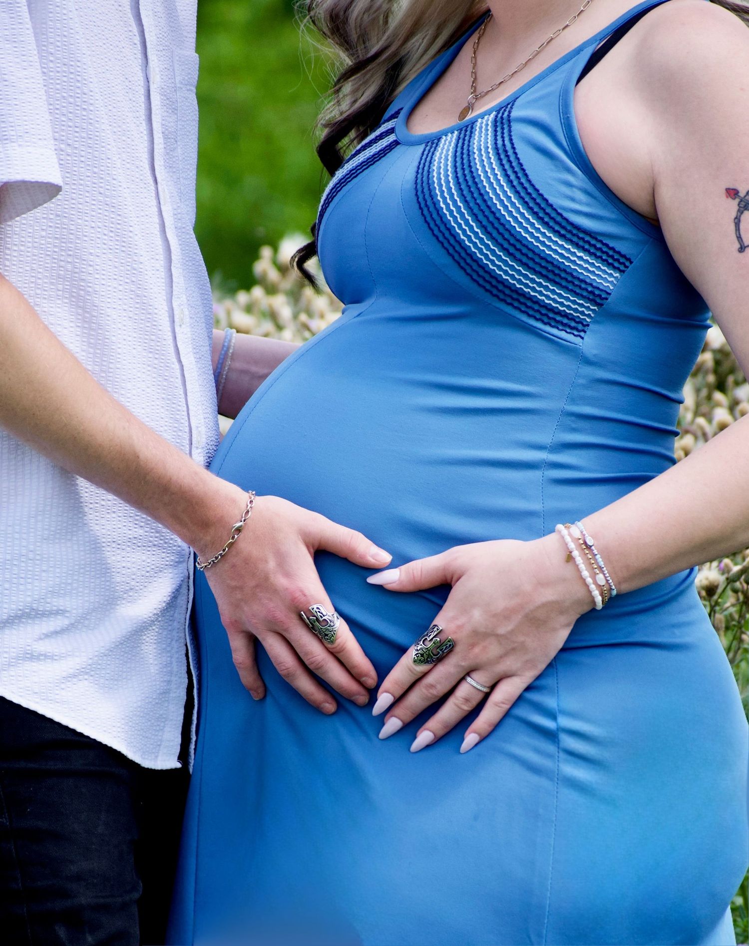 Pregnant woman in blue dress with partner both cradling baby bump outdoors, maternity photo session.