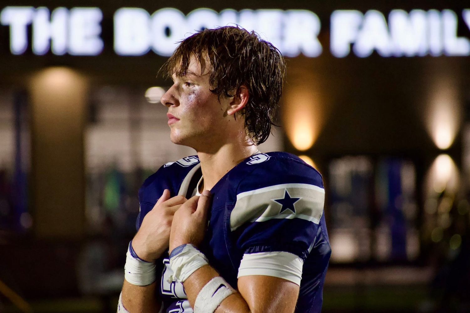 Young football player in blue uniform standing reflectively under stadium lights after game.