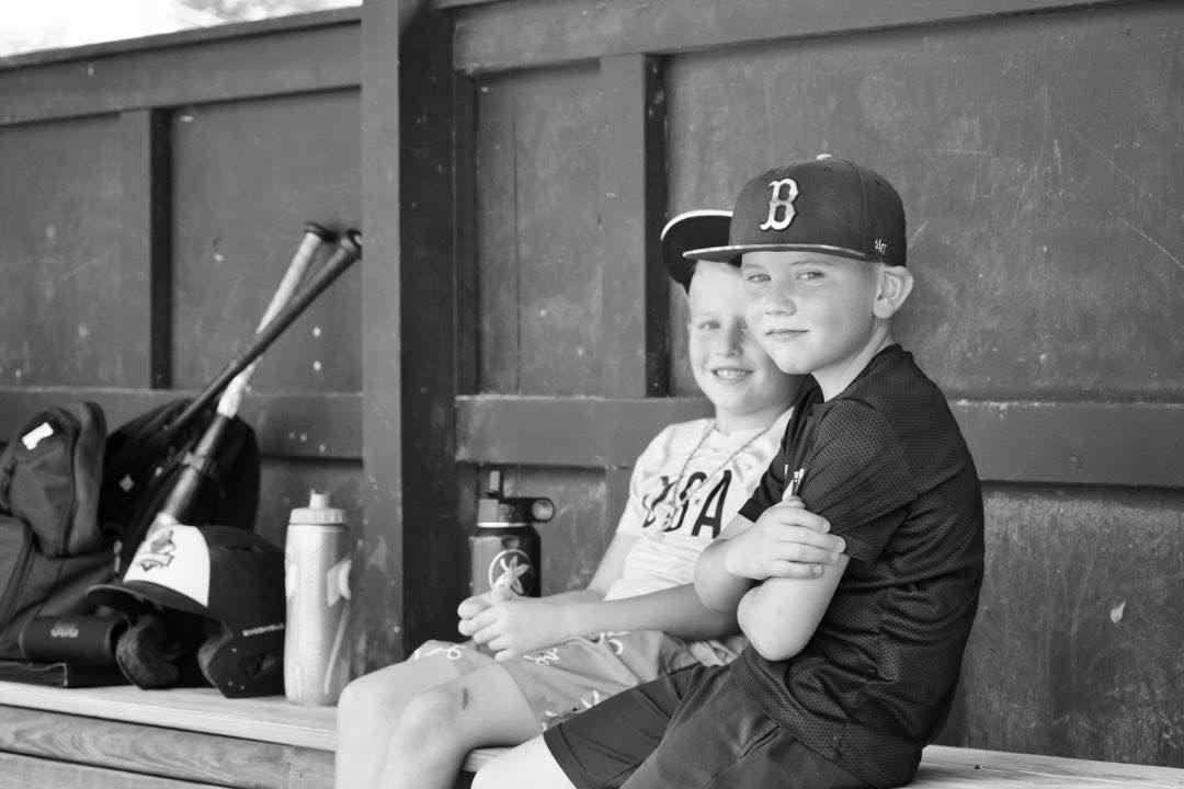 Two young boys in baseball uniforms smiling together in a dugout with bats and gear in black and white.