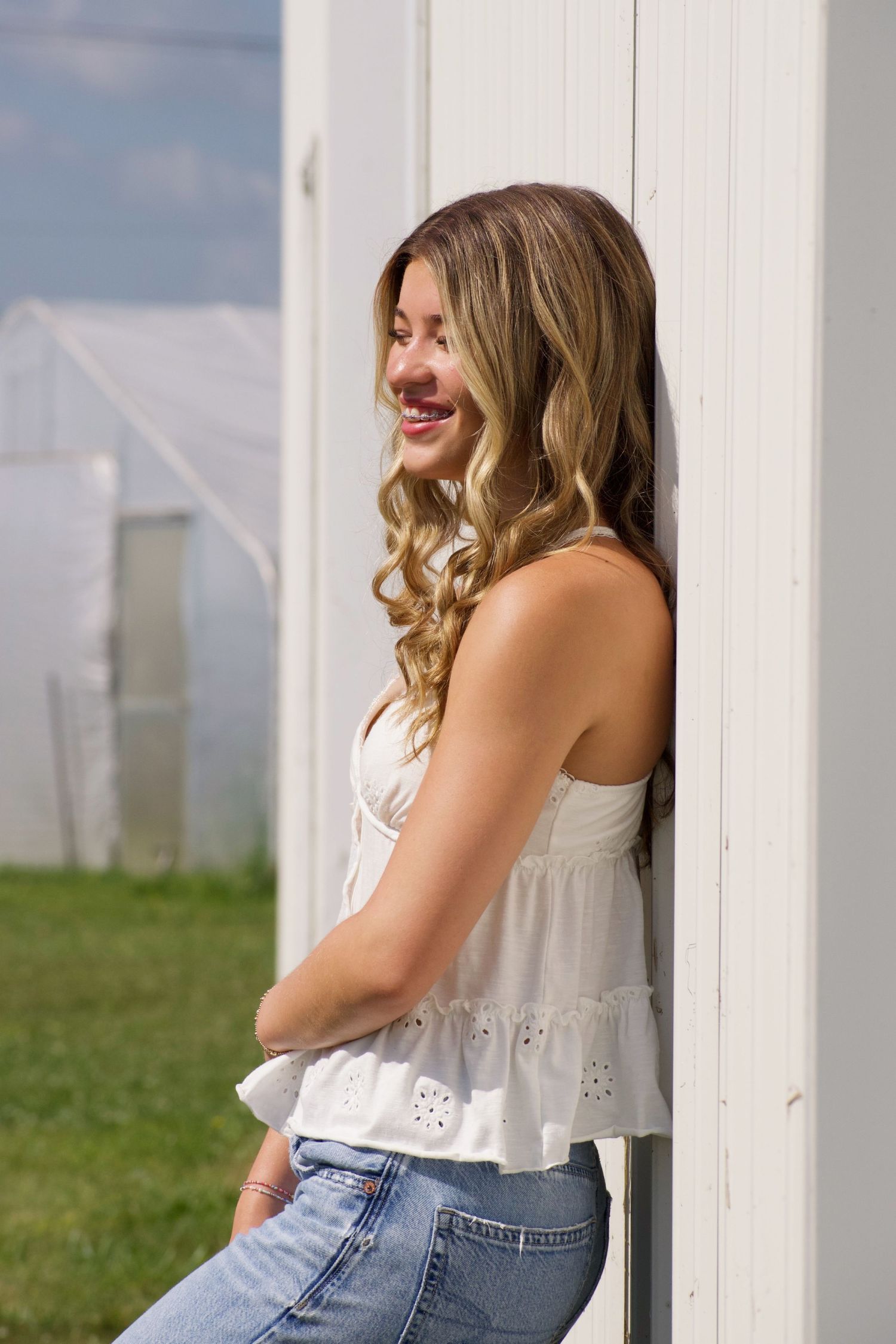 Young woman in white top and jeans leaning against white wall outdoors smiling.