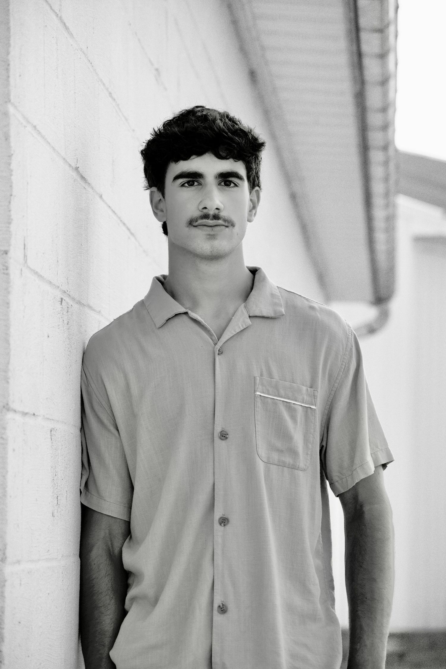Young man with mustache in button-up shirt leaning against white brick wall, B&W photo.