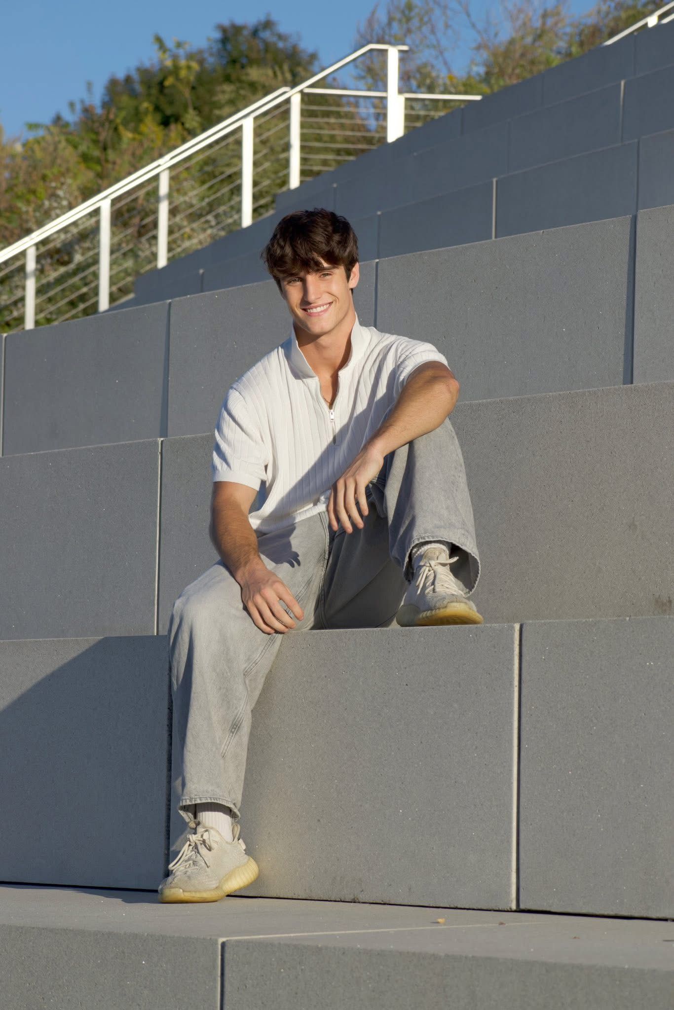 Young man in white polo shirt sitting on gray concrete steps outdoors smiling.