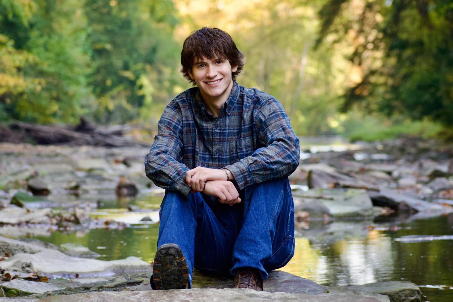 Smiling teenage boy crouching on rocks beside a stream in autumn forest setting.