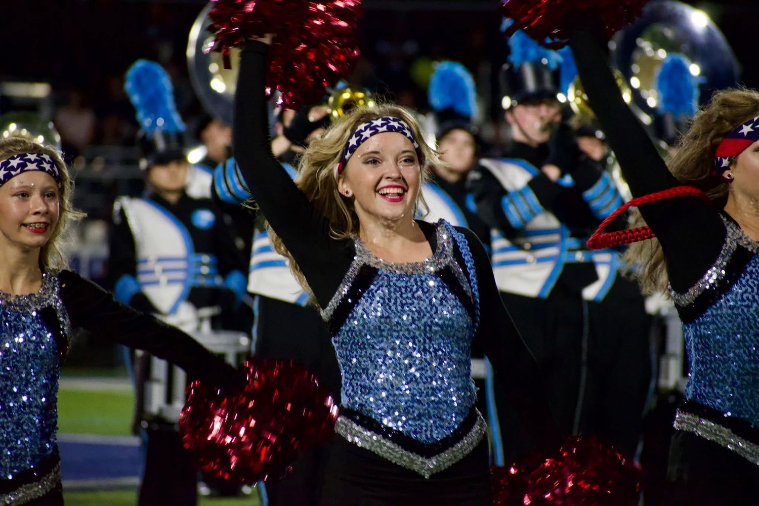 Cheerleader in sparkly blue costume with red pom-poms performing at a night football game.