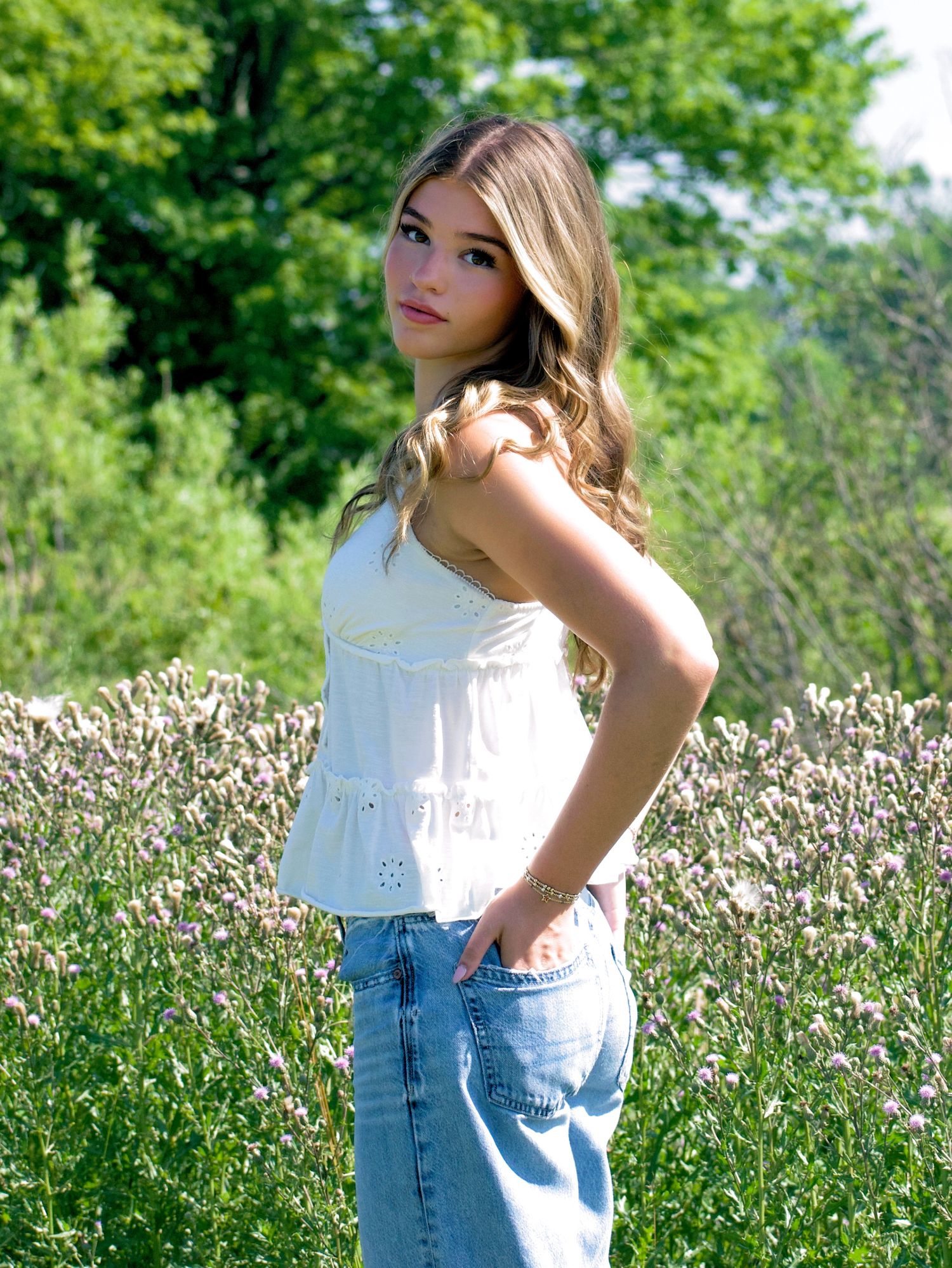 Teen girl in white eyelet top and jeans posing in wildflower field, side profile.