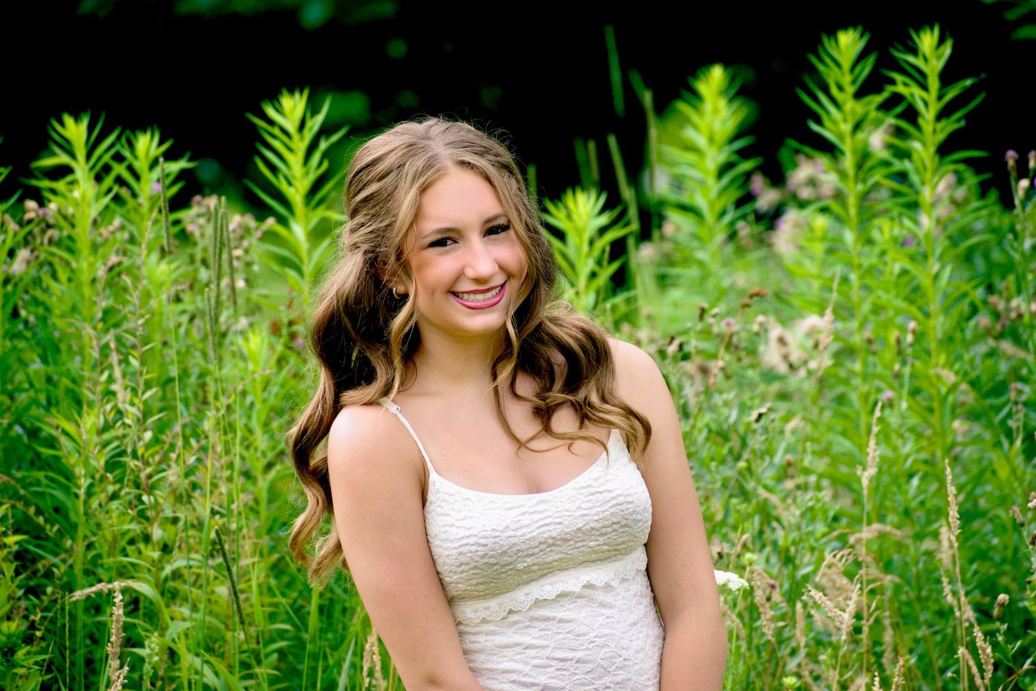 Young woman in white dress smiling outdoors among tall green plants in a lush garden setting.