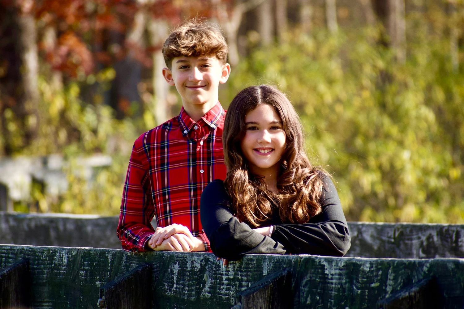 Two smiling siblings, a boy and girl, leaning on a rustic fence in autumn outdoor setting.