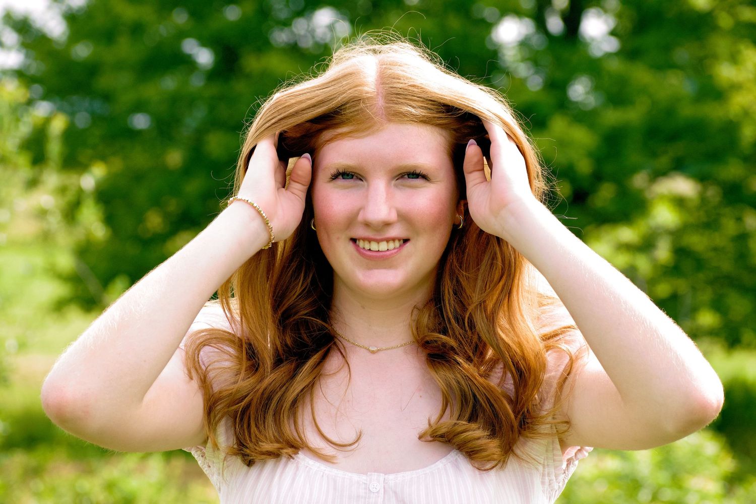 Smiling young redhead woman with freckles holding her hair back outdoors with green trees behind her.
