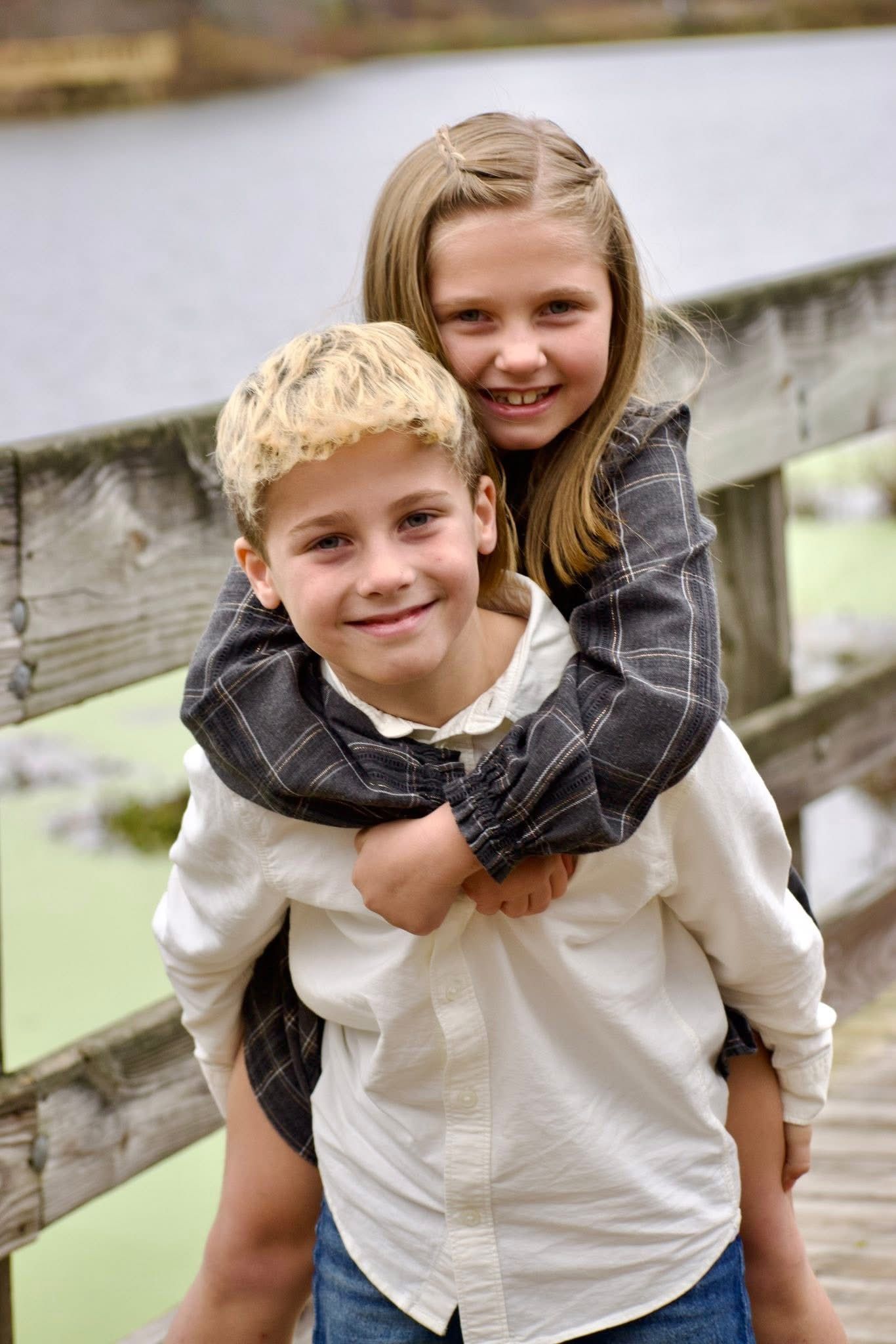 Smiling blonde boy giving piggyback ride to older sister on wooden bridge near pond.