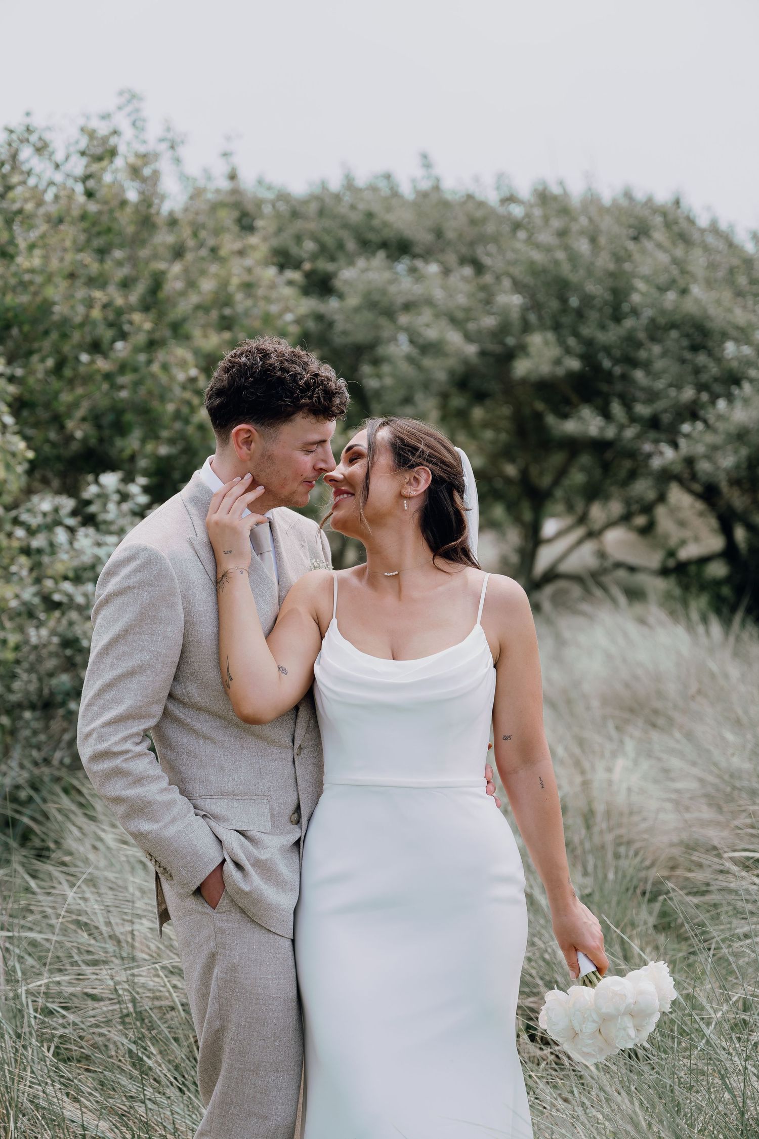 A couple in wedding attire shares a romantic moment outdoors, the bride in white holding a bouquet and groom in a gray suit.