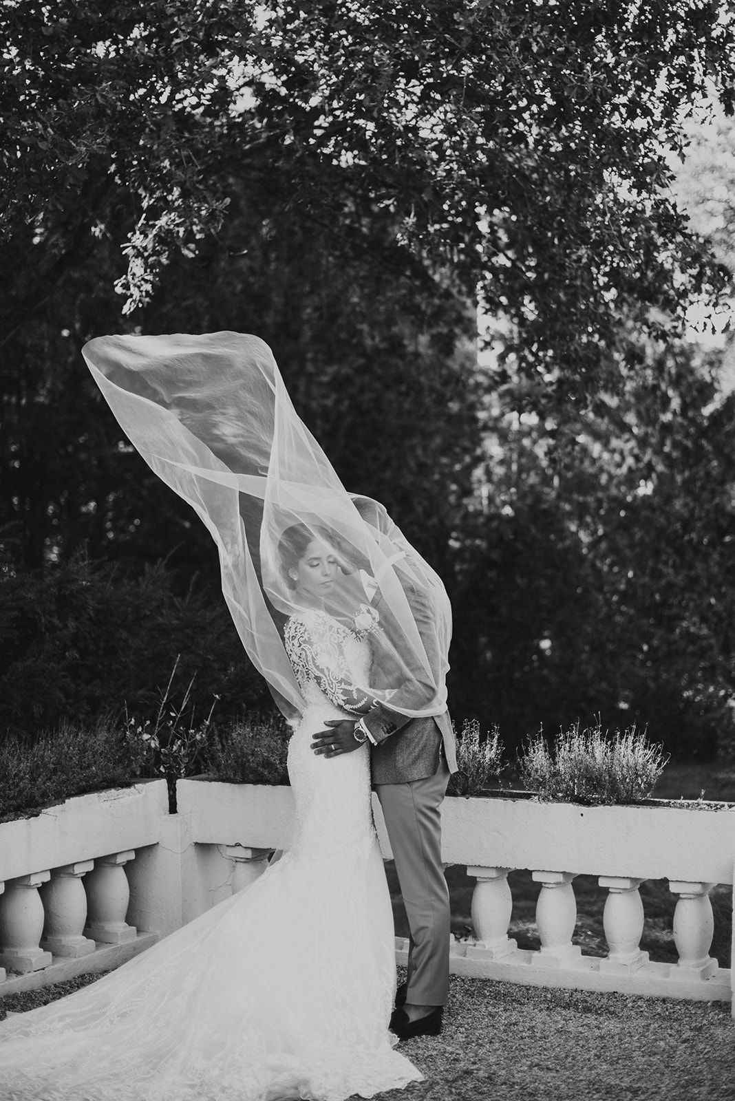 Black and white wedding photo of bride and groom with veil billowing in the wind outdoors.
