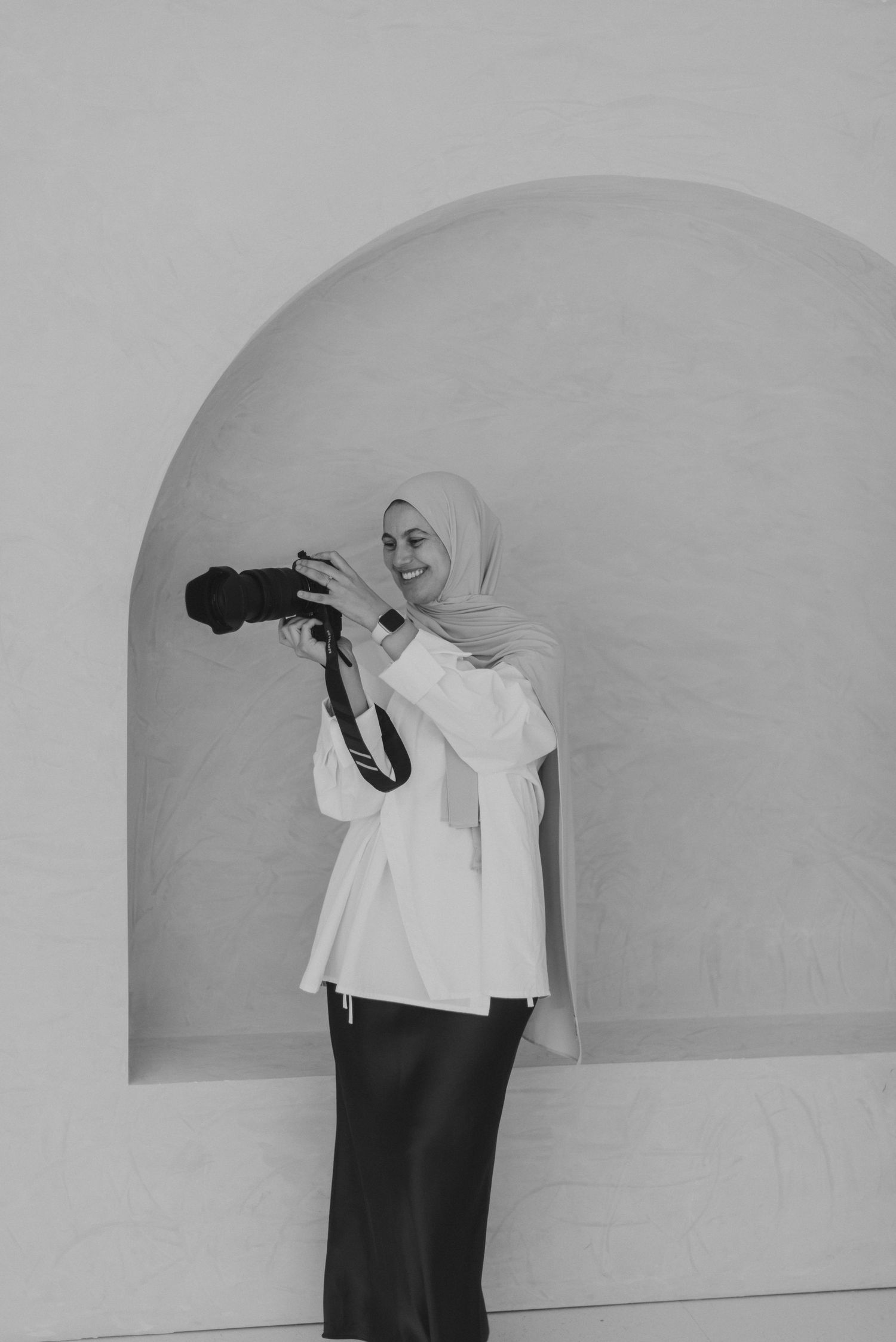 Black and white photo of a smiling woman in hijab and white blazer holding a camera in an arched alcove.