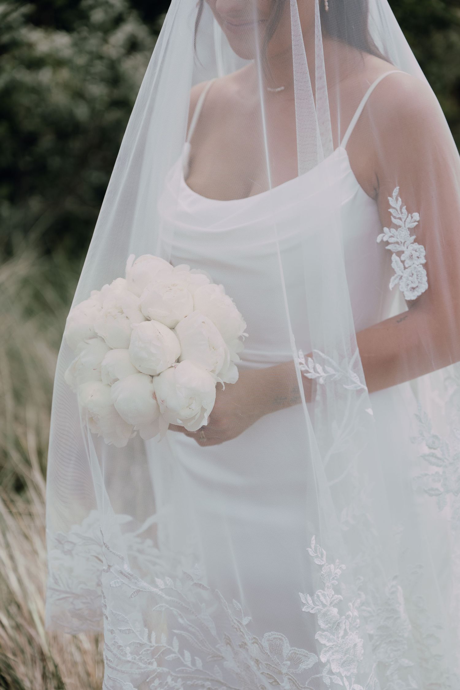 Bride in white slip dress holding white peony bouquet, wearing lace-trimmed veil outdoors.