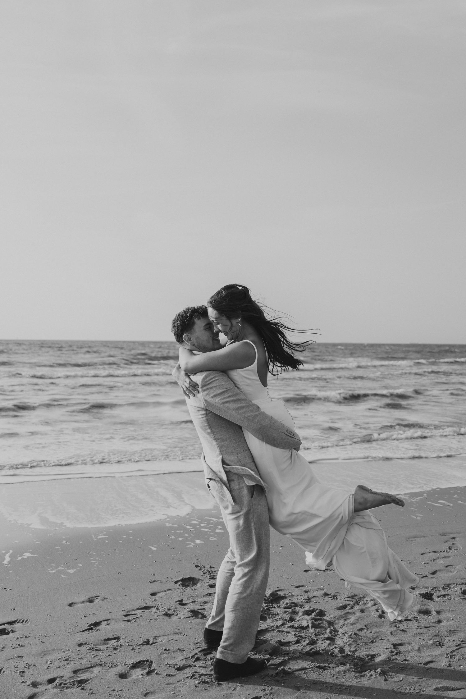 A groom lifts his bride on a beach in a romantic black and white wedding photo.