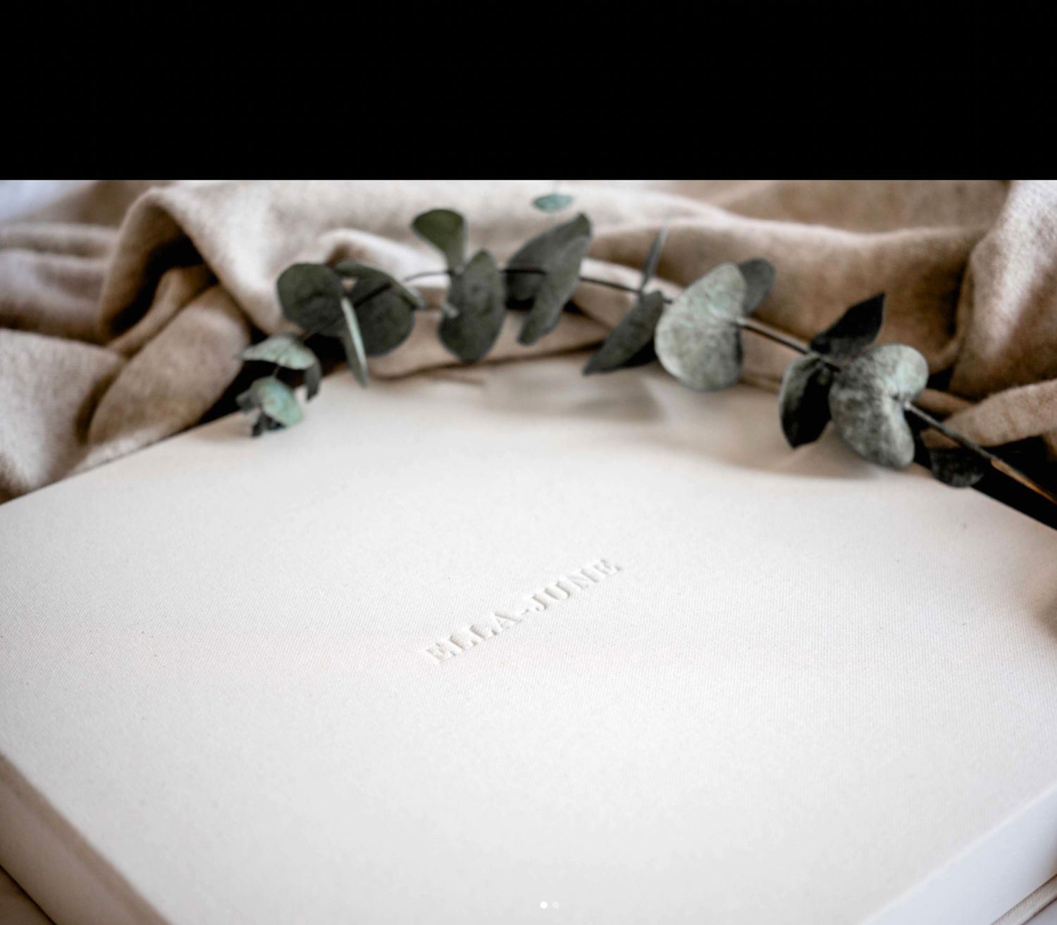 Cream white photo album with embossed text resting on beige fabric with eucalyptus sprigs.