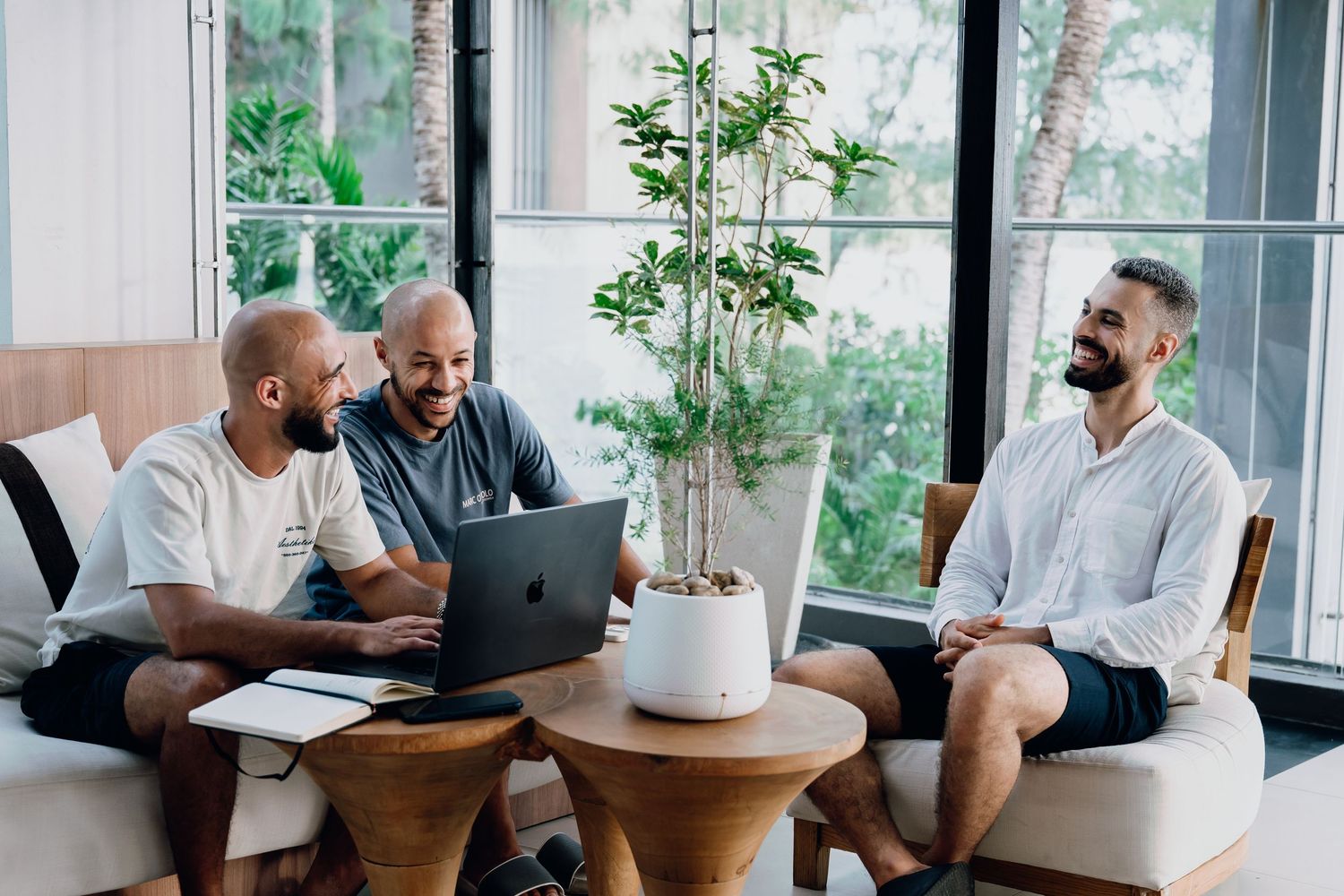 Three men smiling and collaborating on a laptop in a bright modern lounge with tropical plants.