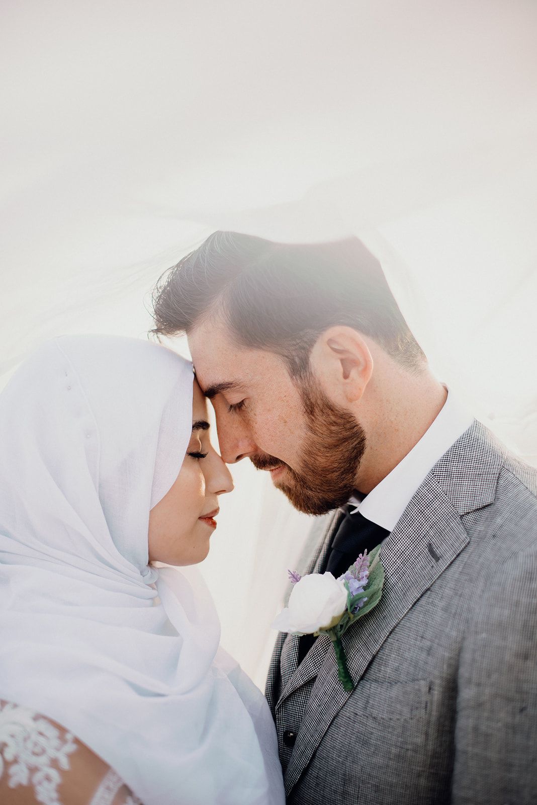 Muslim bride in white hijab and groom in grey suit share an intimate forehead-to-forehead moment.