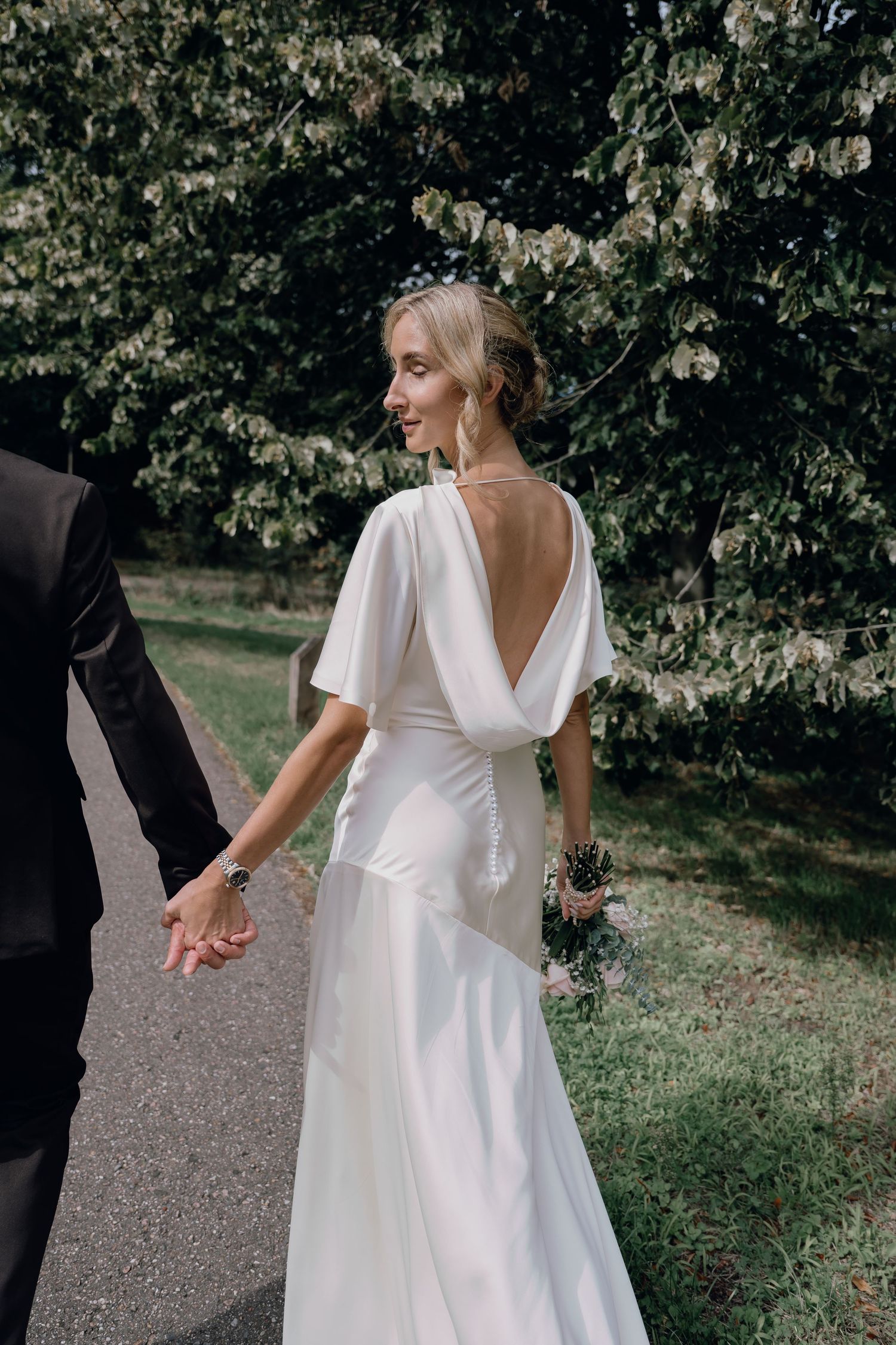 Bride in elegant white backless wedding gown with flutter sleeves holds groom's hand while walking outdoors.