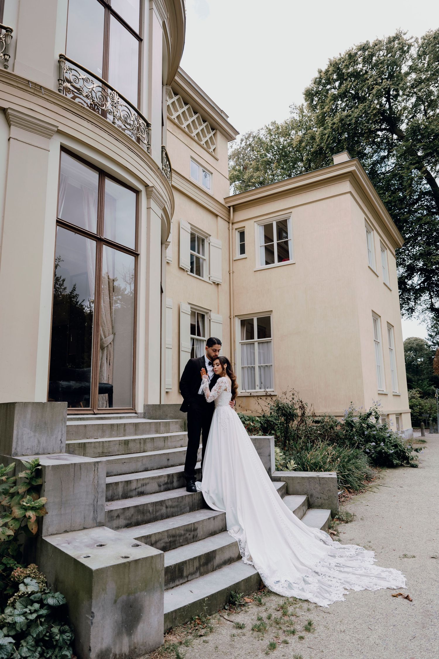Bride and groom embracing on stone steps outside a grand cream-colored estate surrounded by greenery.