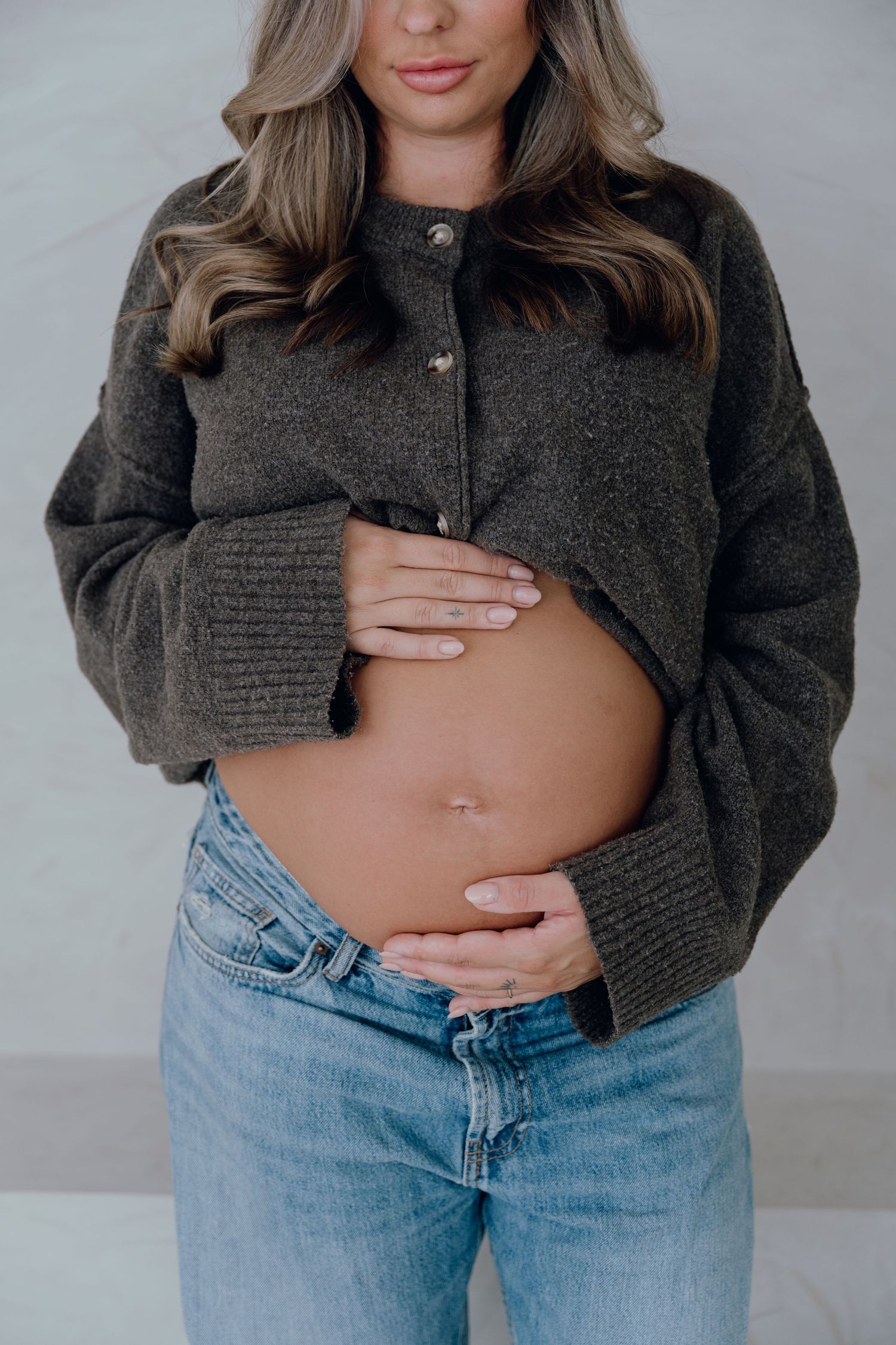 Pregnant woman in dark cropped cardigan and blue jeans cradling her baby bump with both hands.