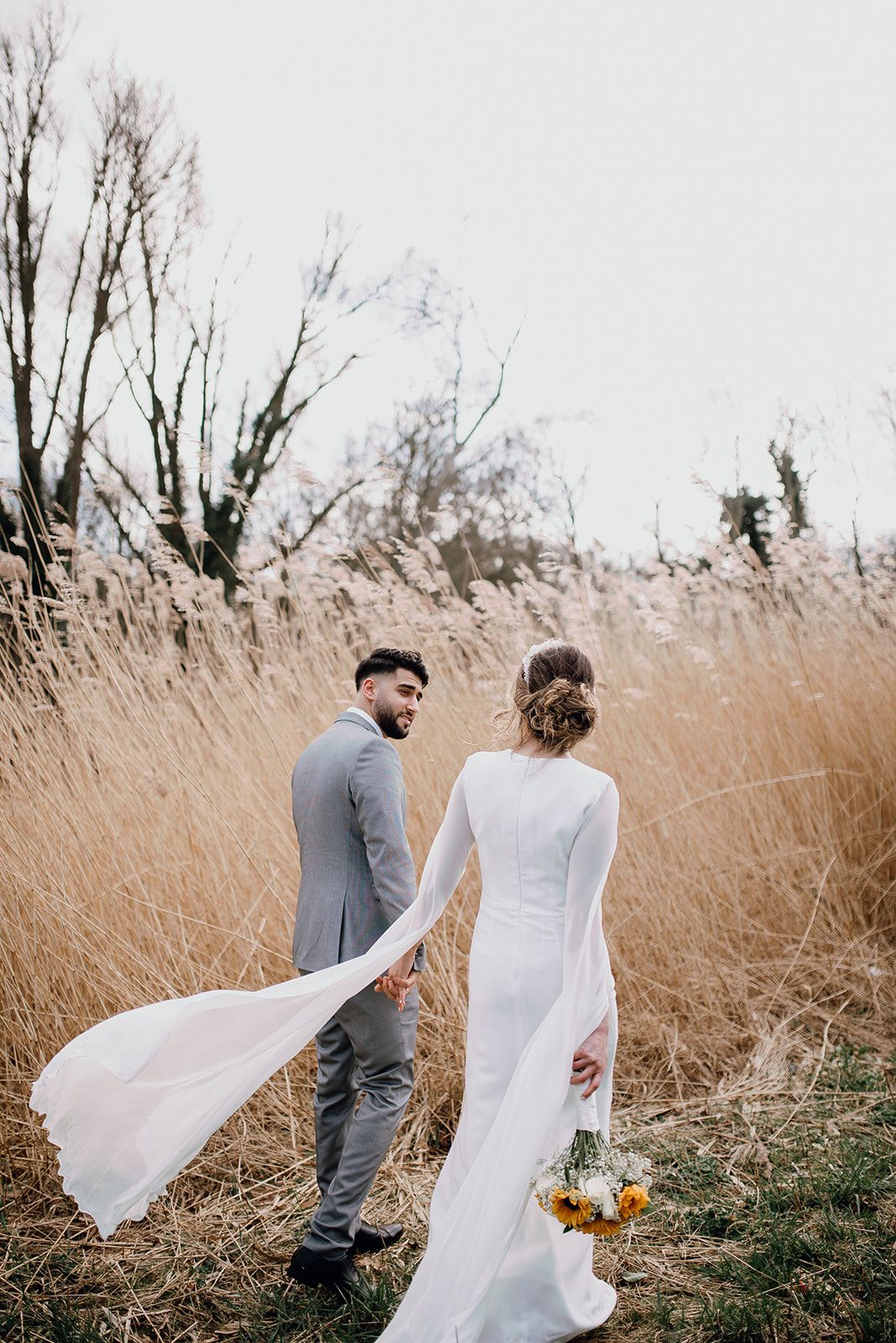 Bride and groom walking through golden wheat field, bride's white gown trailing in the wind on their wedding day.