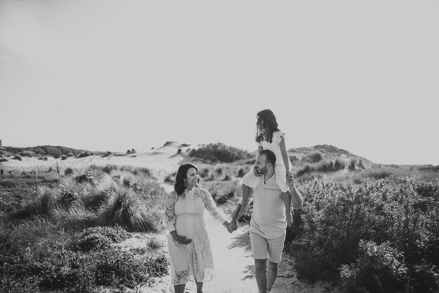 Black and white image of family walking on sandy dunes with child lifted on shoulders.