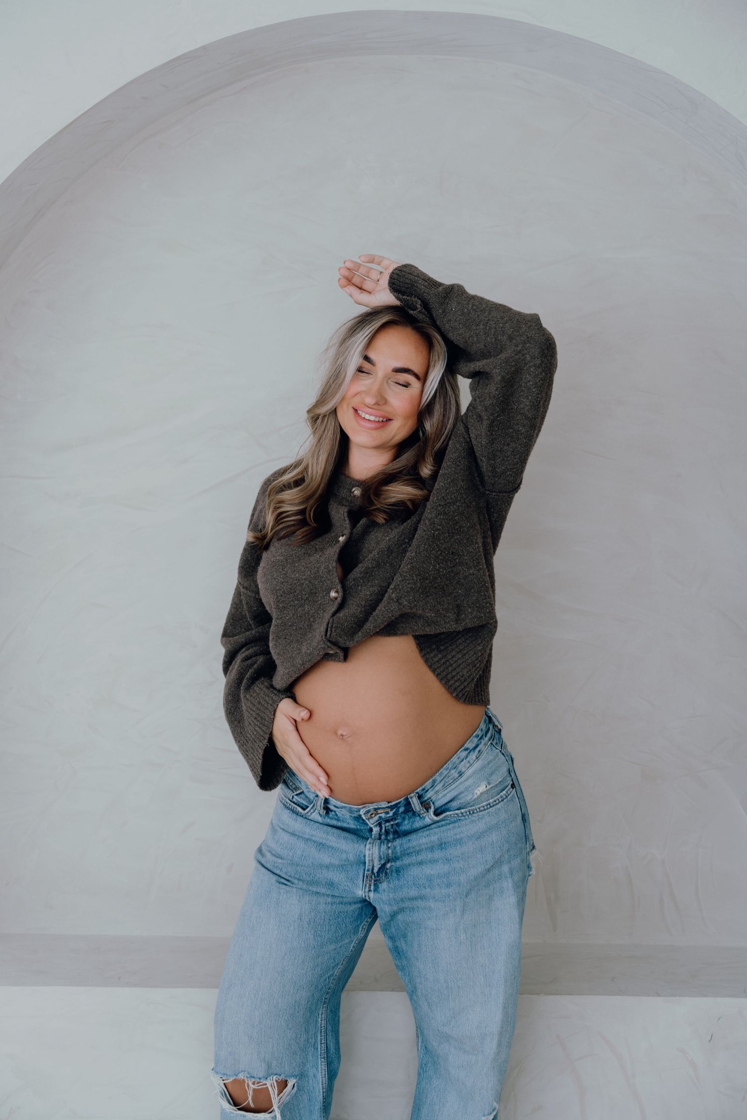 Smiling pregnant woman in gray cropped sweater and ripped jeans posing against white arched wall.