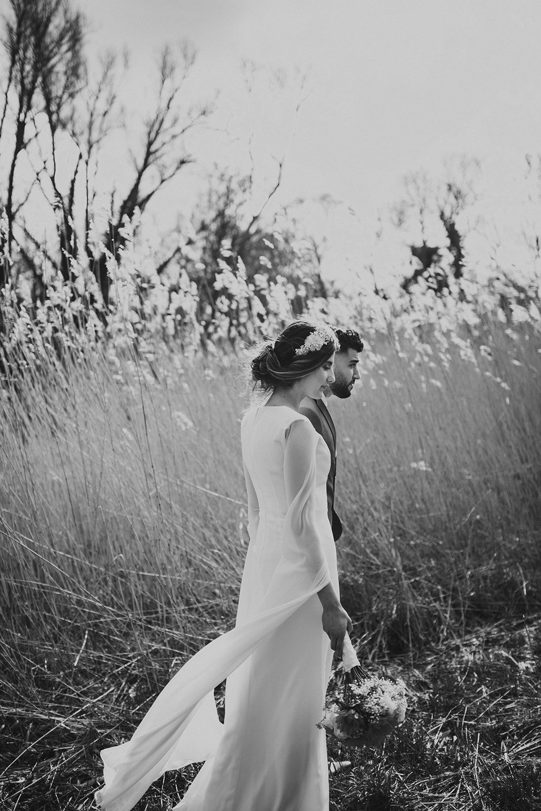 Black and white photo of bride and groom walking through tall grass in natural setting.