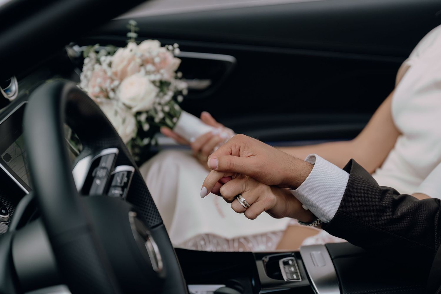 Bride and groom holding hands in a luxury car with a white wedding bouquet on the dashboard.