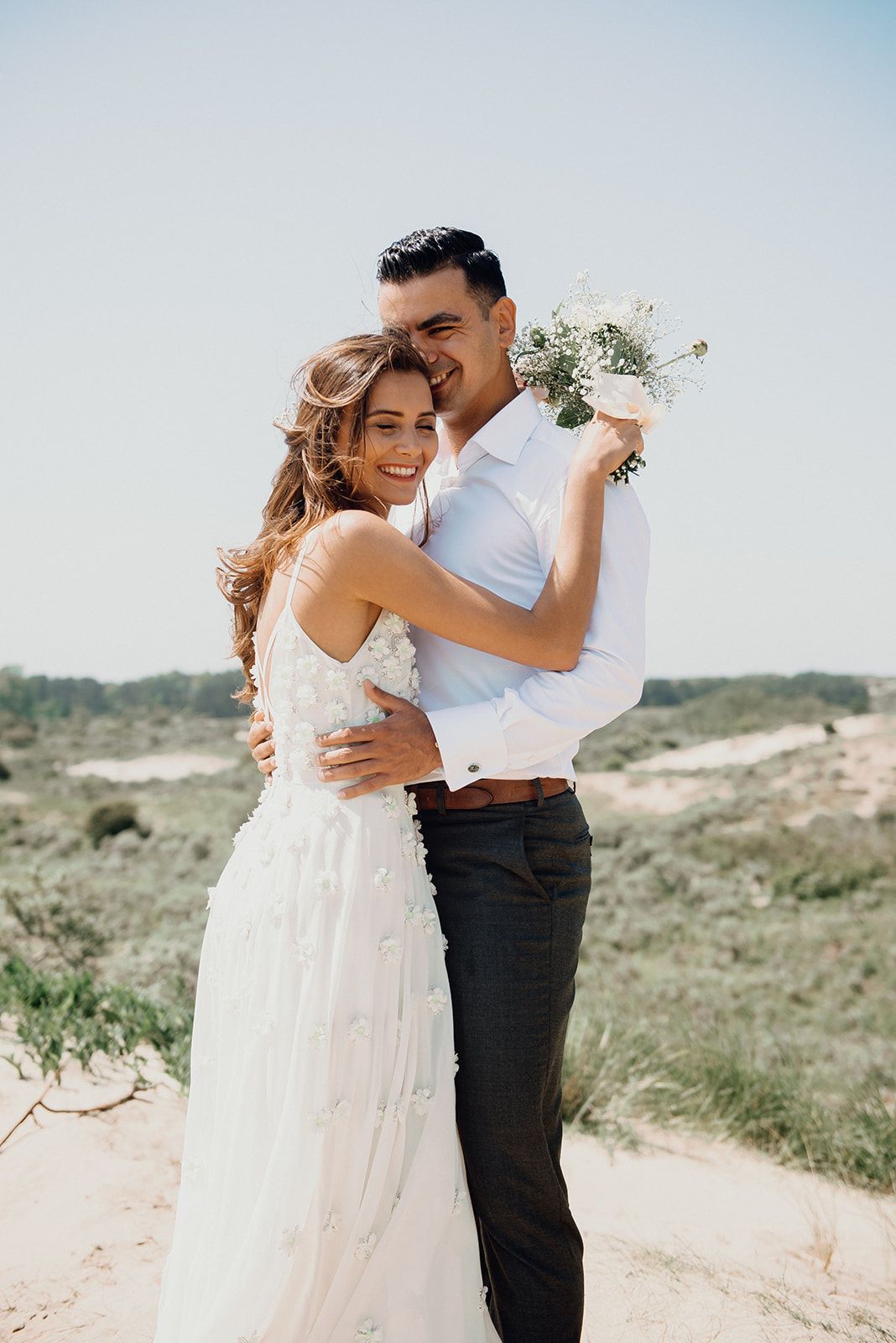 Smiling bride and groom embracing with wildflower bouquet in sandy dunes on sunny day.