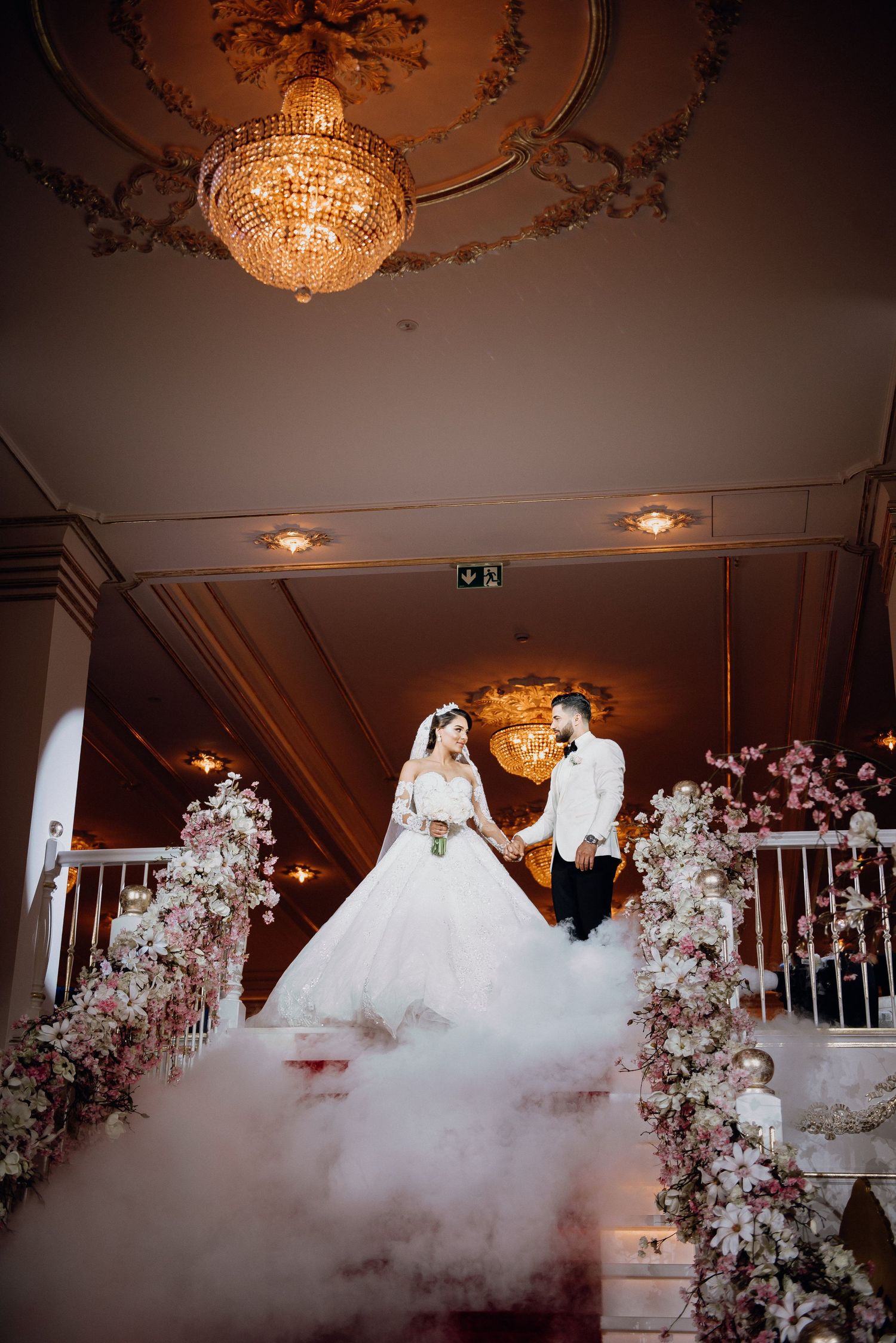 Bride and groom standing on fog-filled staircase surrounded by pink floral arrangements at reception.
