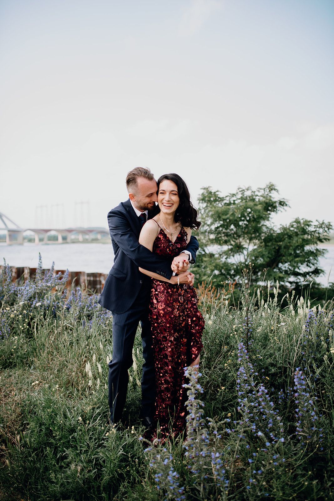 Couple embracing in a wildflower field near a river, woman wearing a red sequin dress.