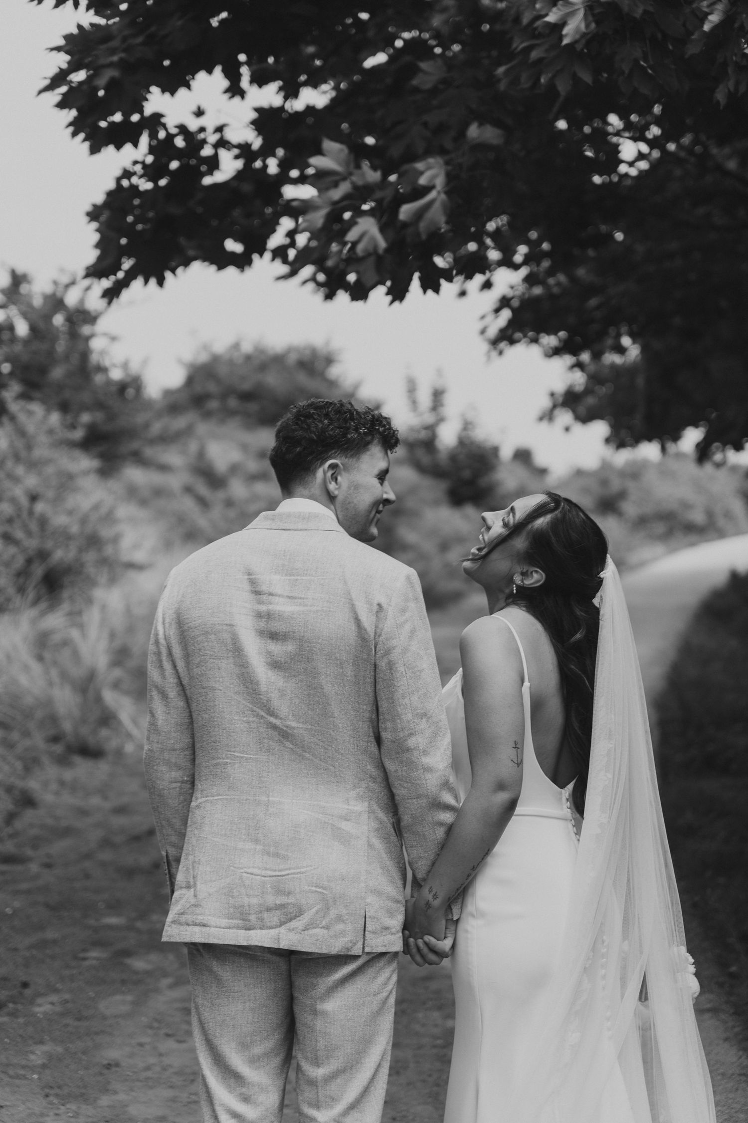 Black and white photo of a bride and groom holding hands, laughing together on a scenic outdoor path.