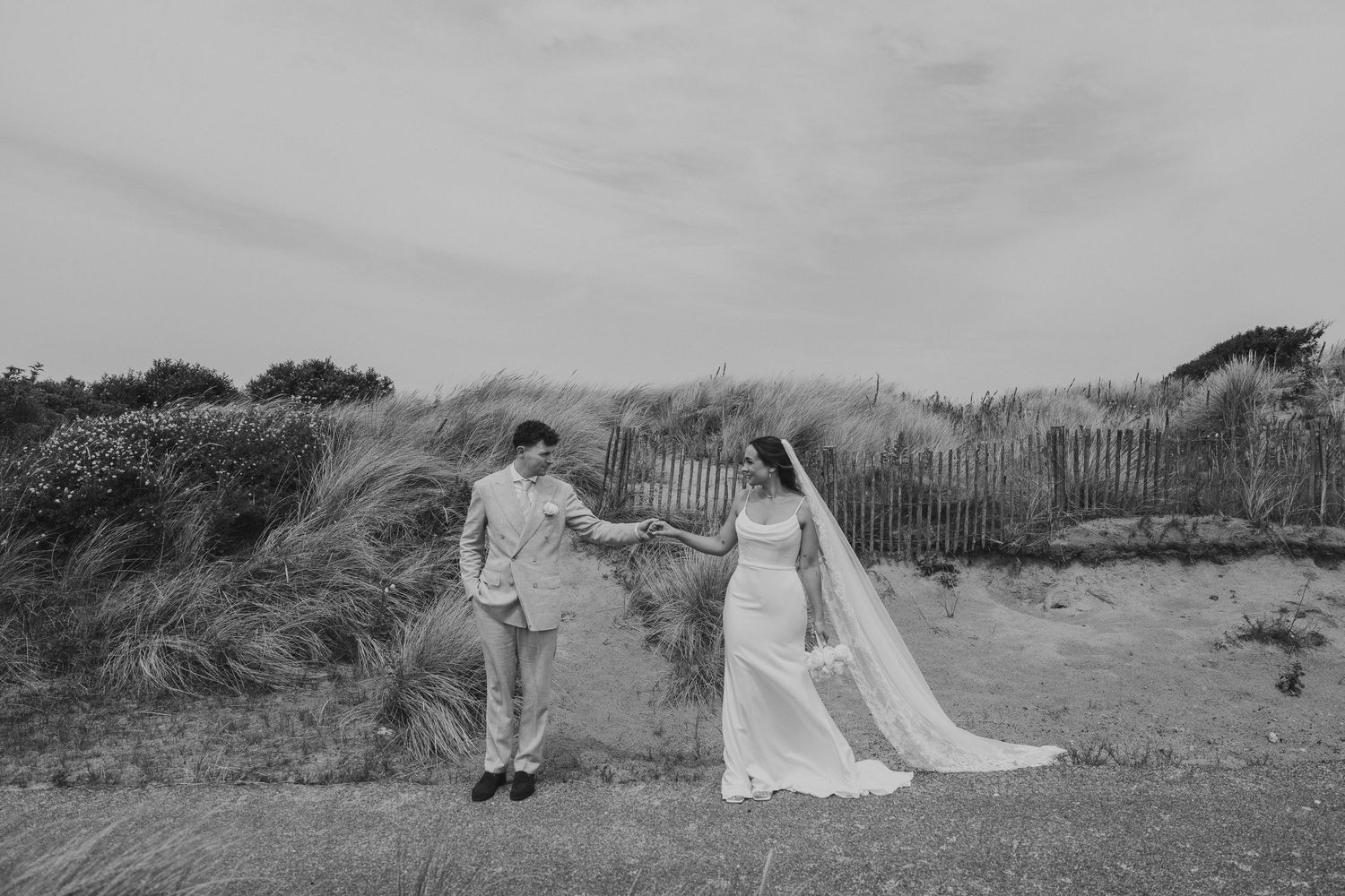 Black and white photo of bride and groom holding hands on coastal dunes with rustic fence backdrop.