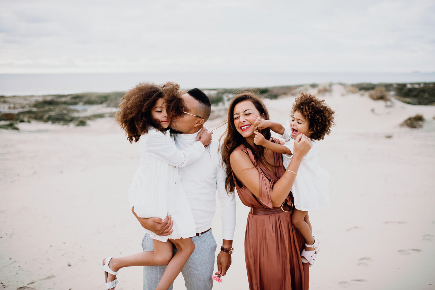 Happy family of four laughing and playing together on a sandy beach on cloudy day.