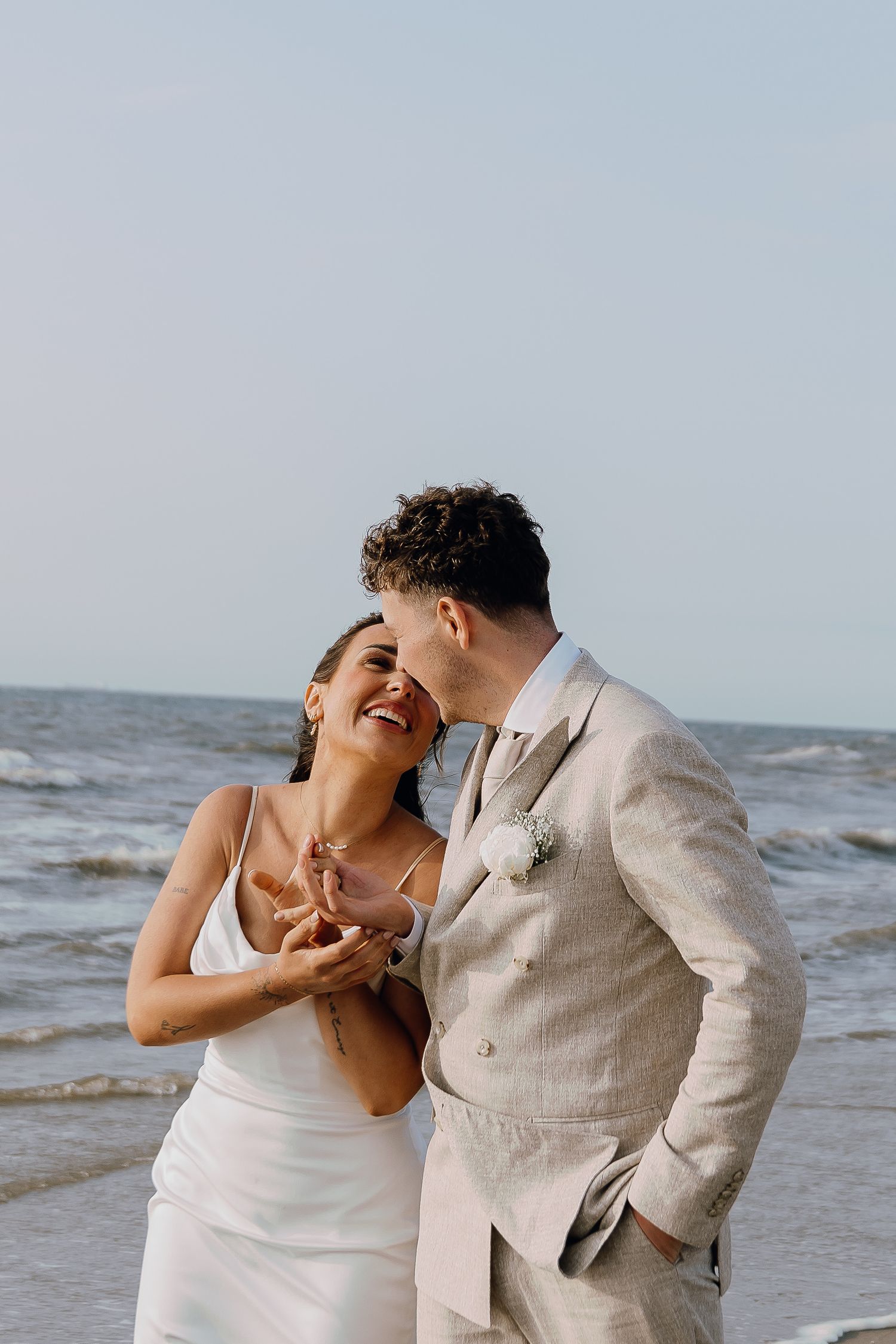 A joyful couple in wedding attire embraces on a beach, the groom kissing the bride's cheek as waves roll behind them.