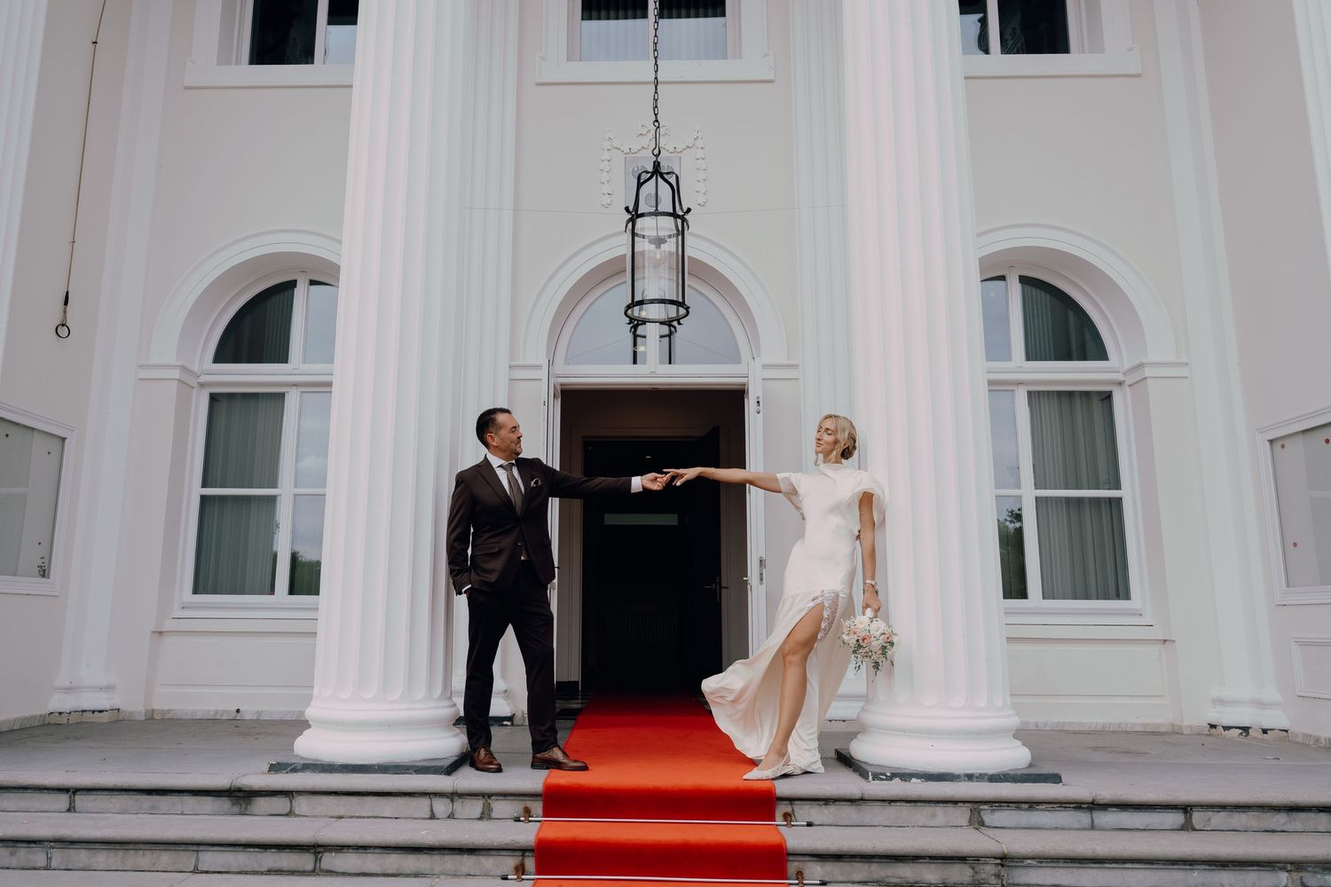 Bride and groom pose on red carpet steps of elegant white building with columns on their wedding day.