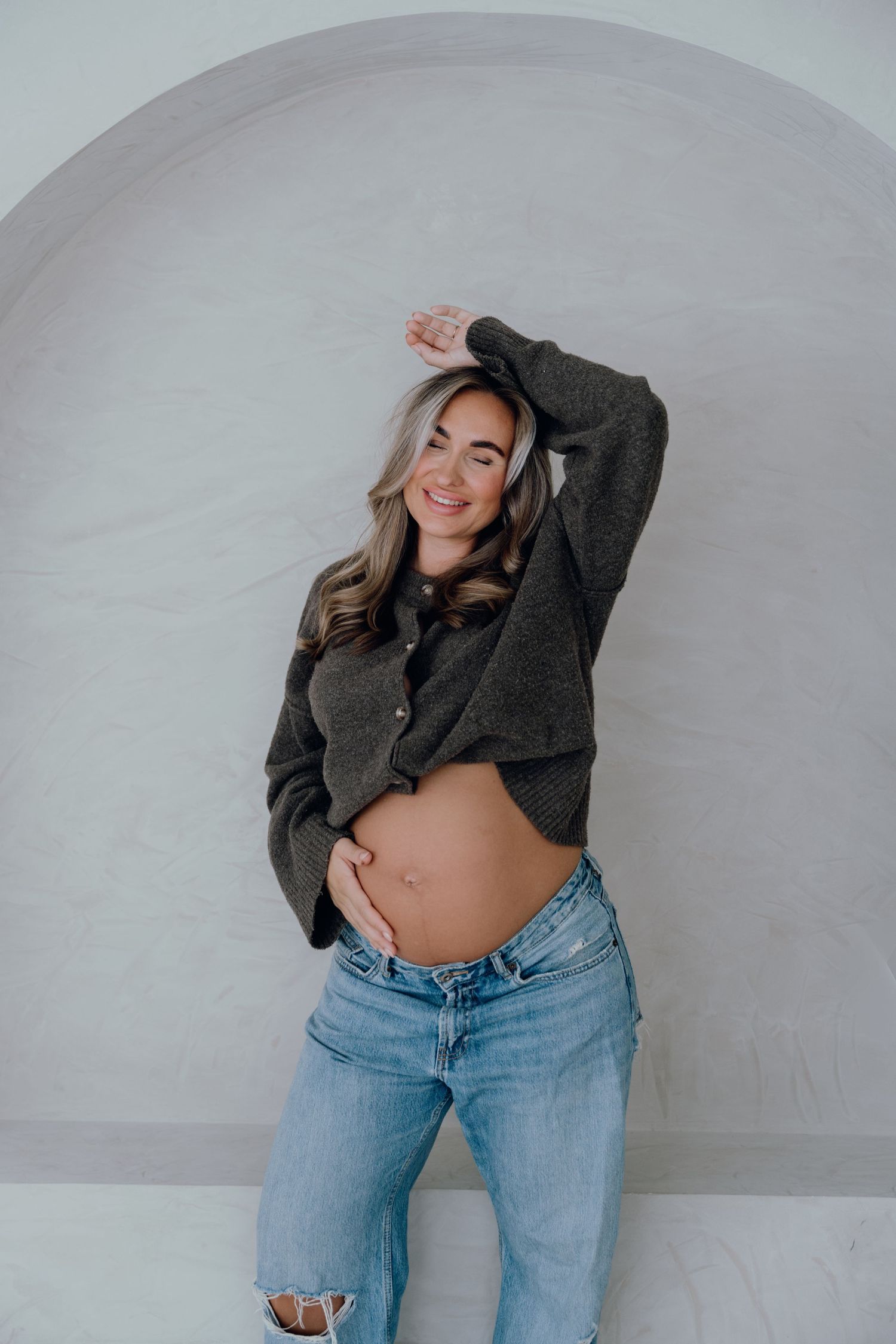 Smiling pregnant woman in gray cropped cardigan and ripped jeans posing against white arched wall.