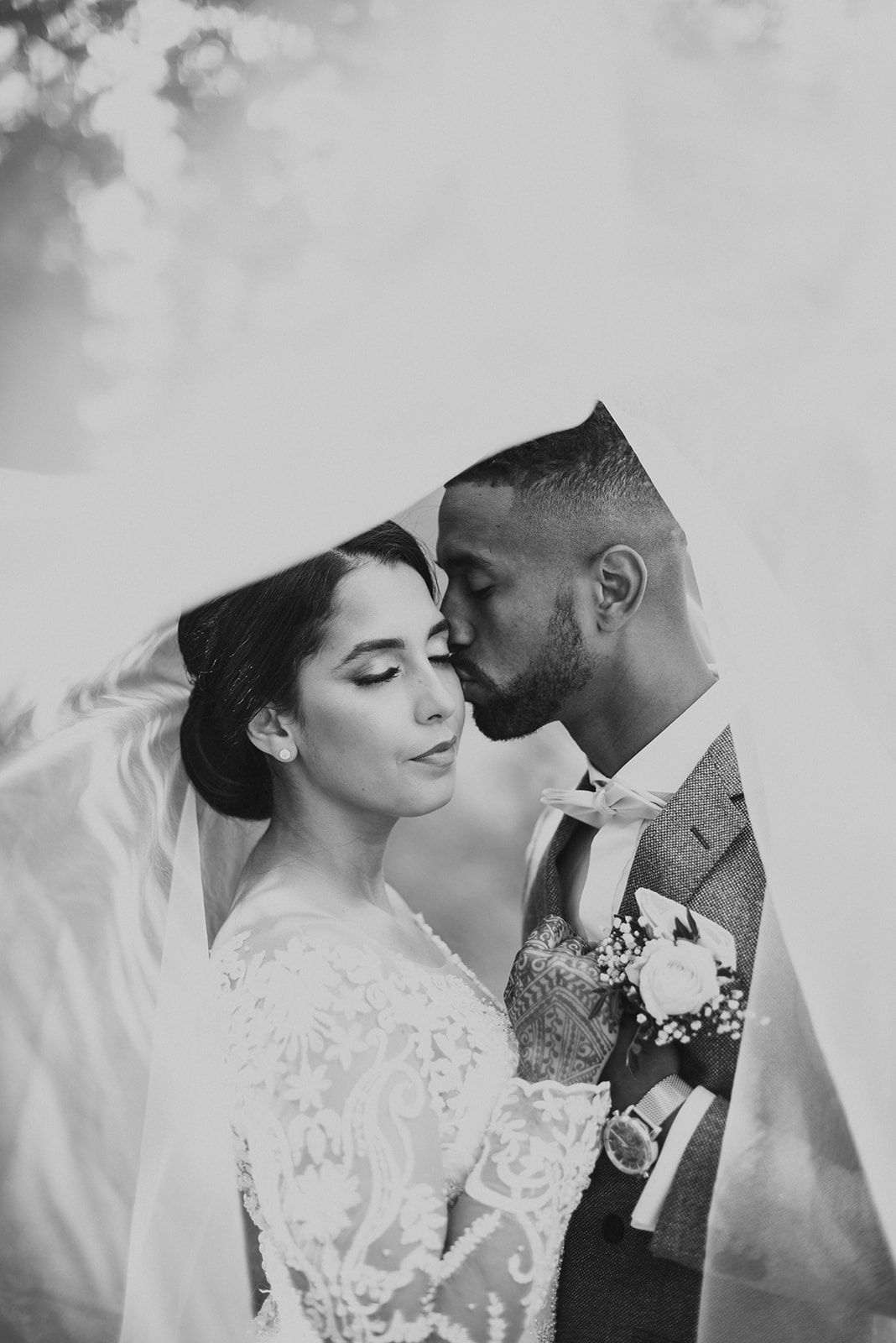 Black and white romantic photo of an interracial couple kissing under a flowing bridal veil.
