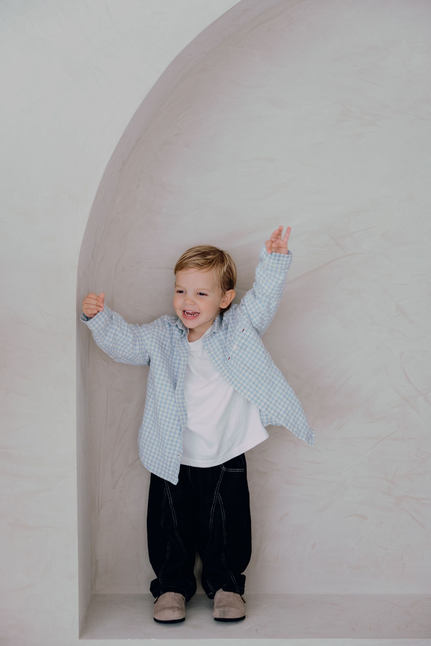 Happy toddler boy in blue plaid shirt and dark pants stands in arched white wall niche with arms raised.