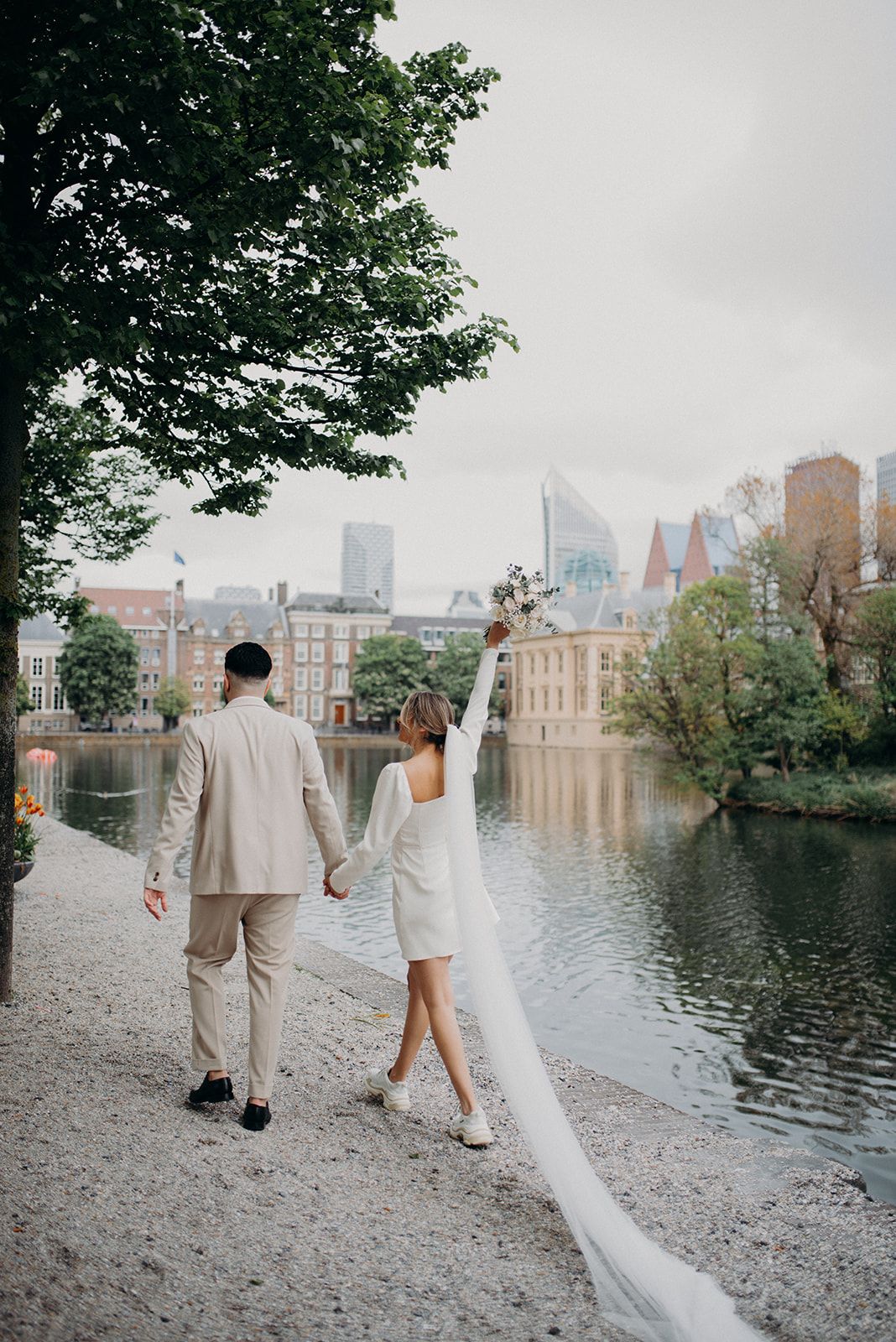 Bride raises bouquet joyfully as couple walks hand in hand along waterfront in The Hague, Netherlands.
