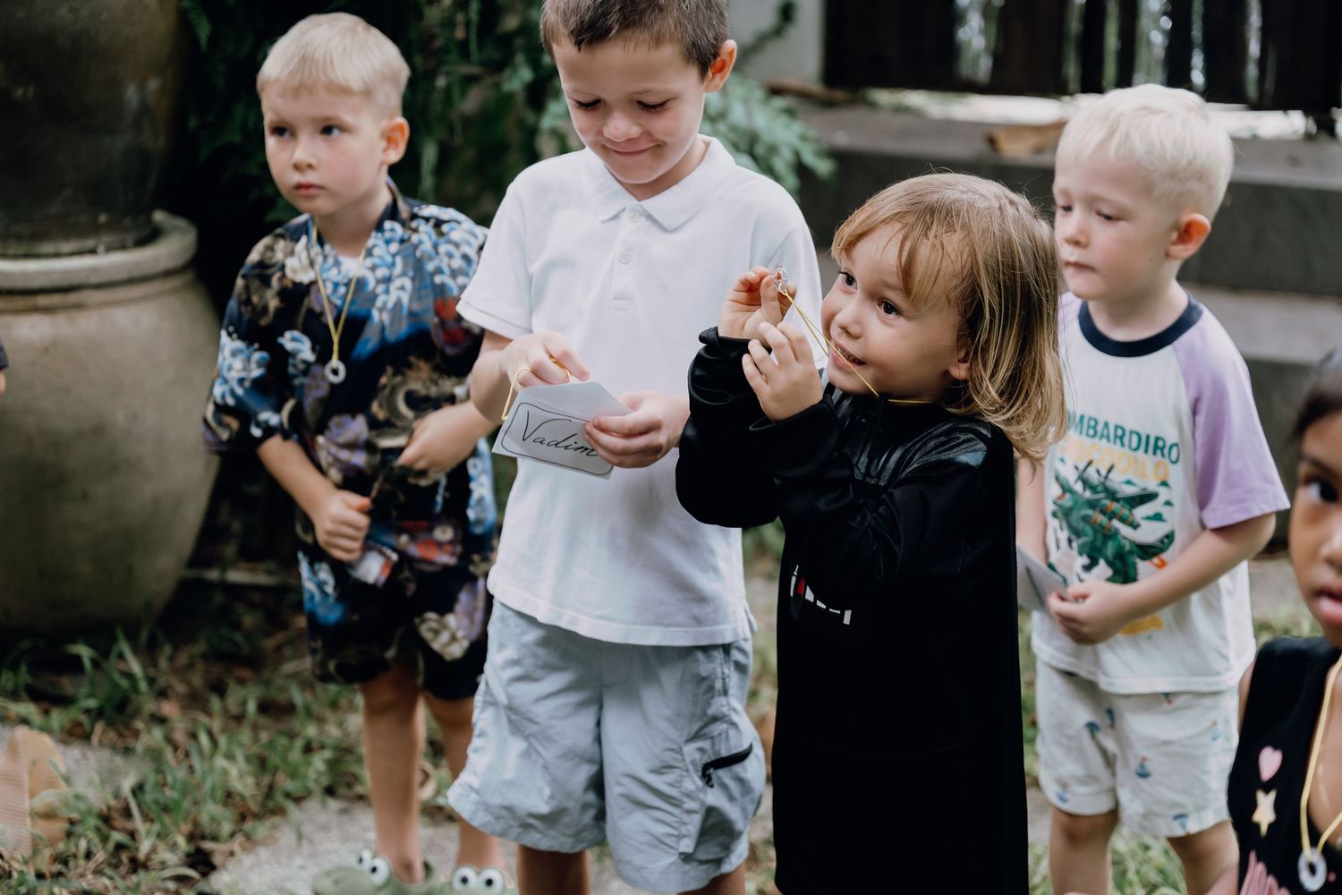 Four young children standing outdoors, examining small objects in their hands at a garden party.