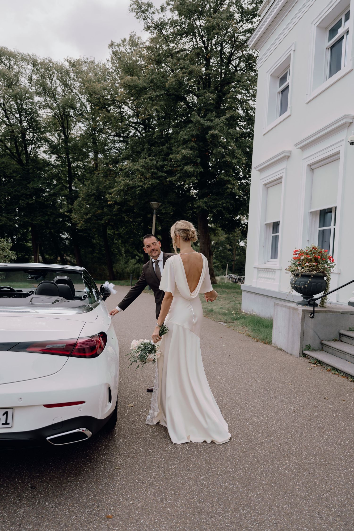 Bride and groom holding hands beside a white convertible car outside an elegant white mansion on their wedding day.