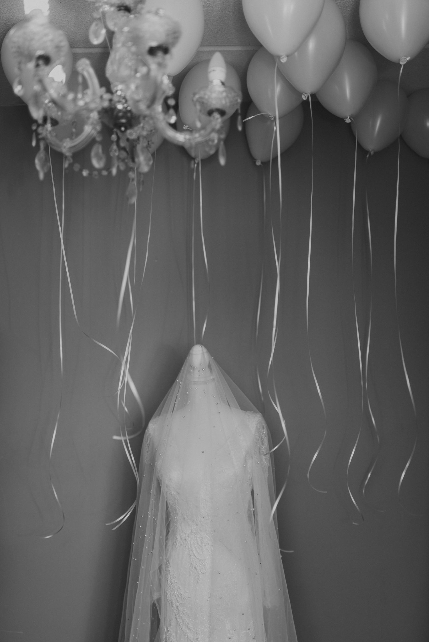 Black and white image of a bride in veil standing beneath floating balloons and a chandelier.