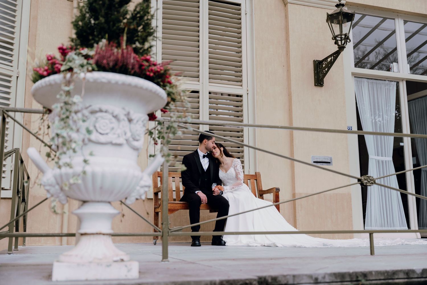 Bride and groom sit together on steps of a grand estate with ornate white urns and floral arrangements.