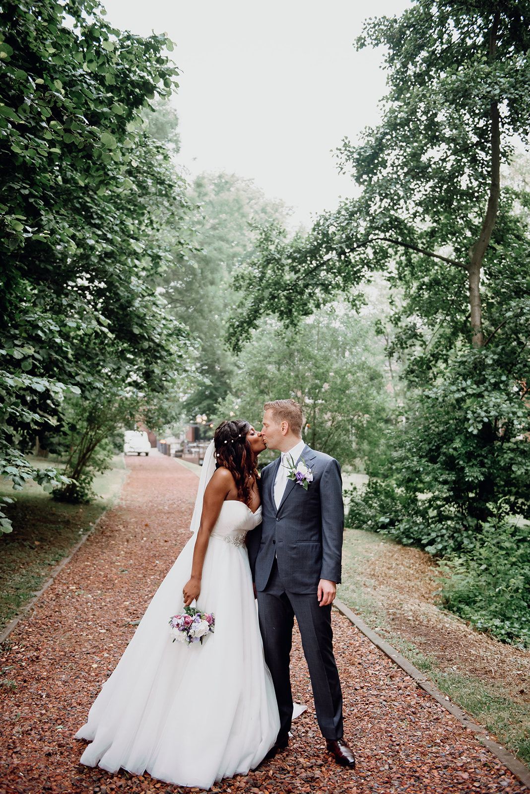 Bride and groom share a kiss on a tree-lined path, bride holding a pink floral bouquet.