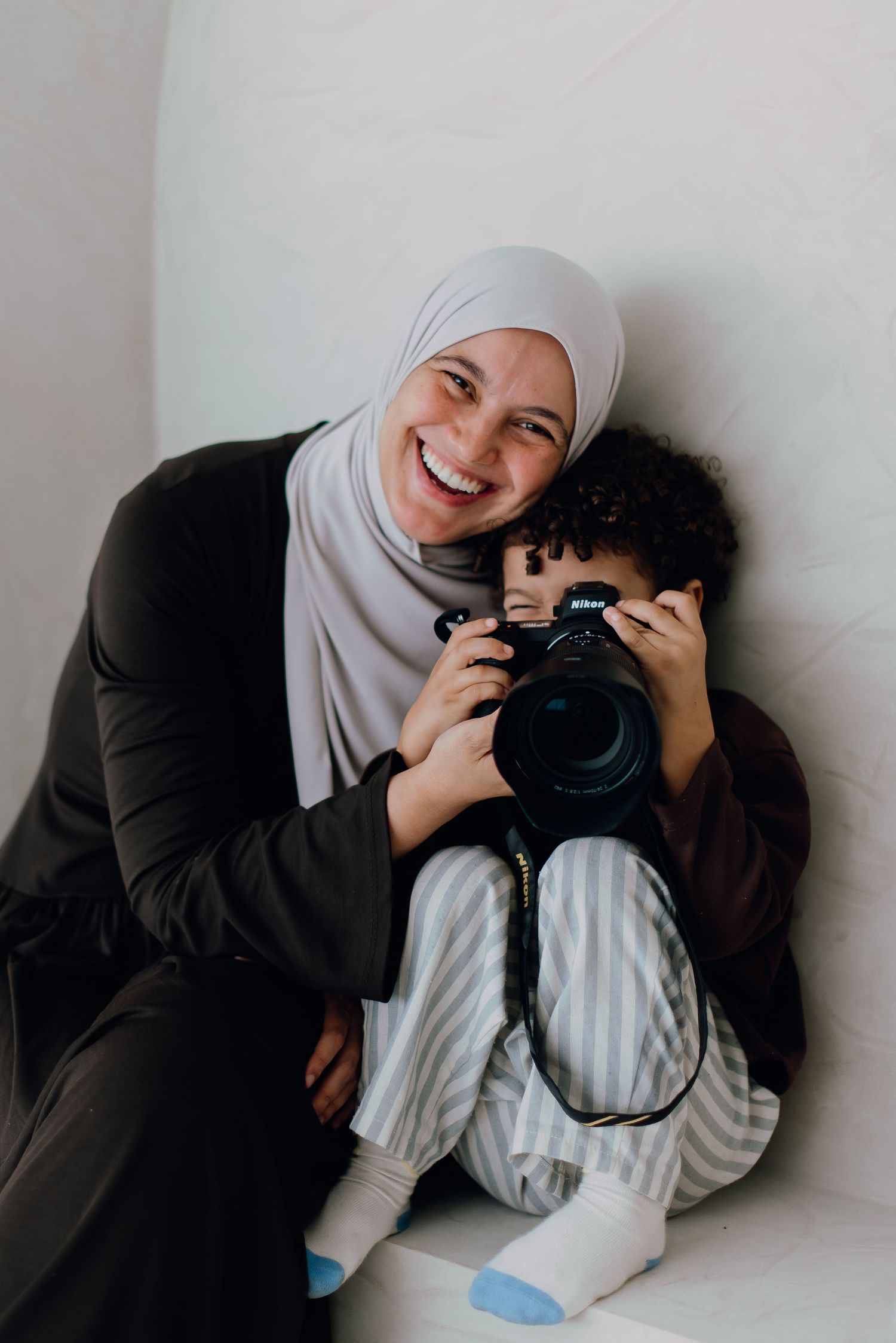 A smiling woman in a hijab hugs a child holding a Nikon camera while sitting against a white wall.