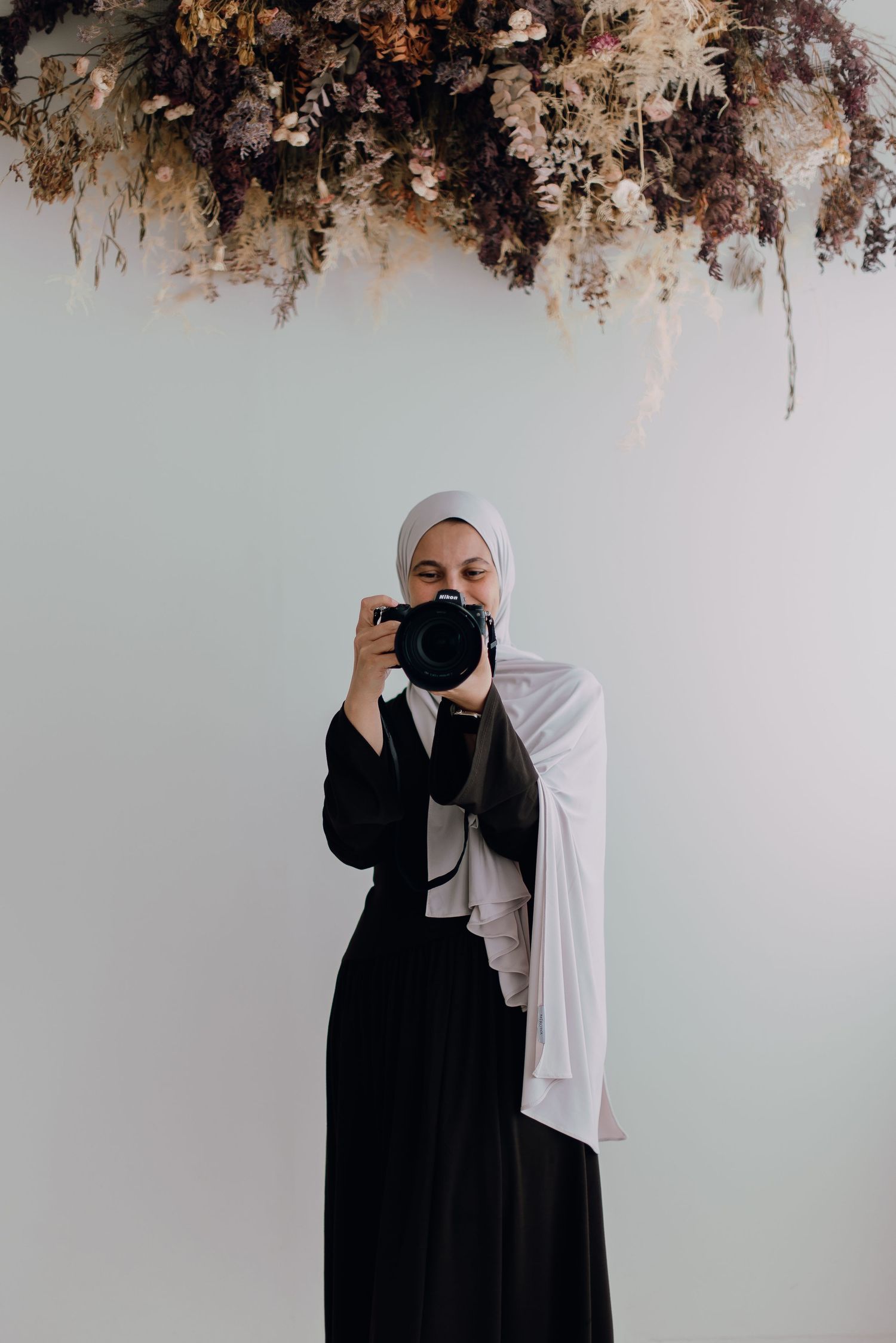 A woman in a black abaya and white hijab holds a DSLR camera up to her face beneath a dried floral installation.