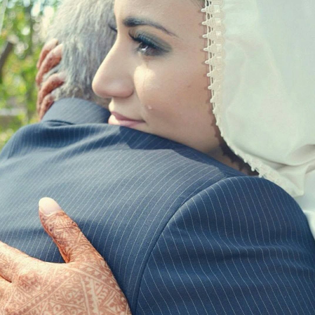Emotional bride in white hijab with henna hands embracing someone in a suit, shedding tears of joy.