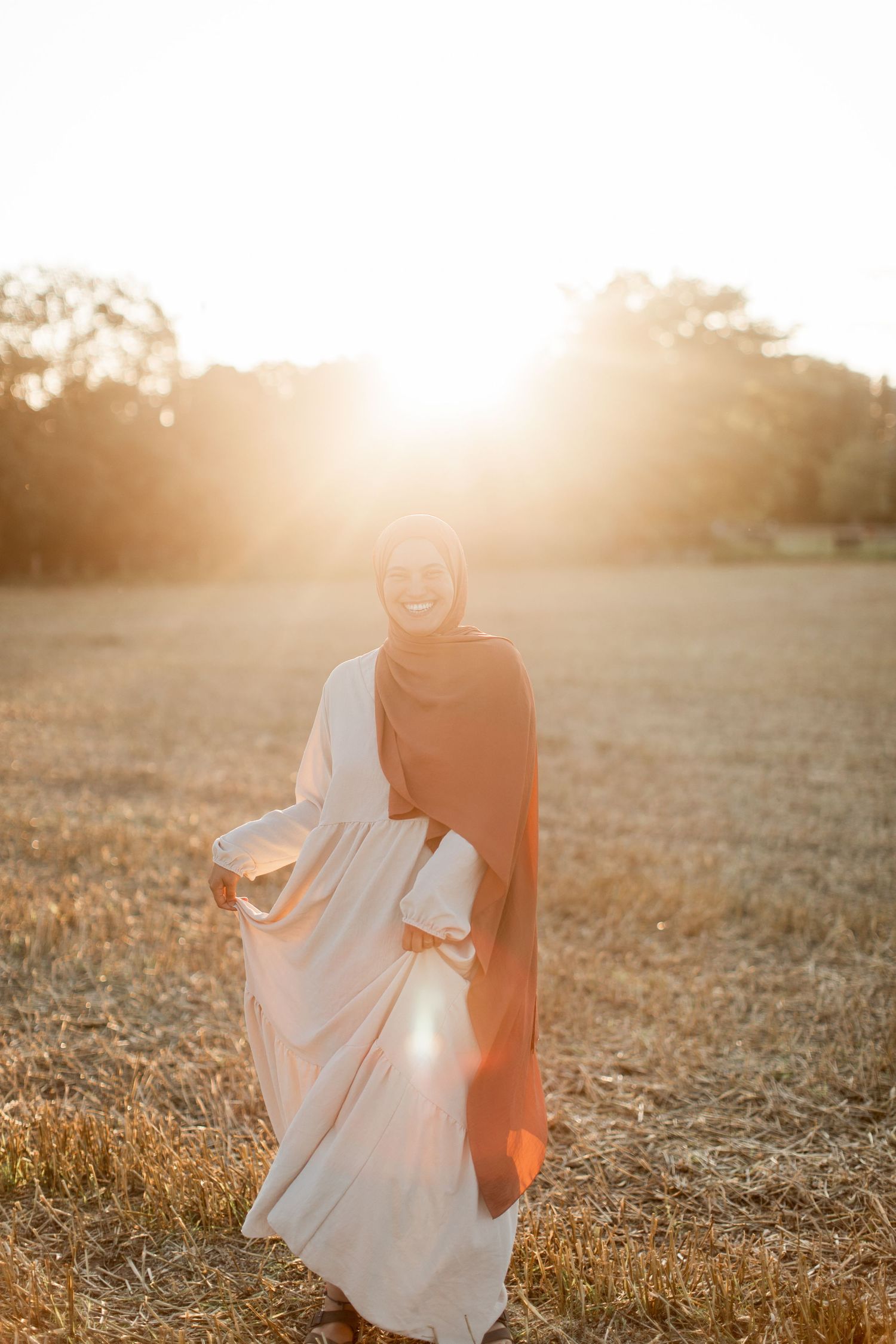 A smiling woman in a hijab and flowing dress stands in a golden field during a stunning sunset.